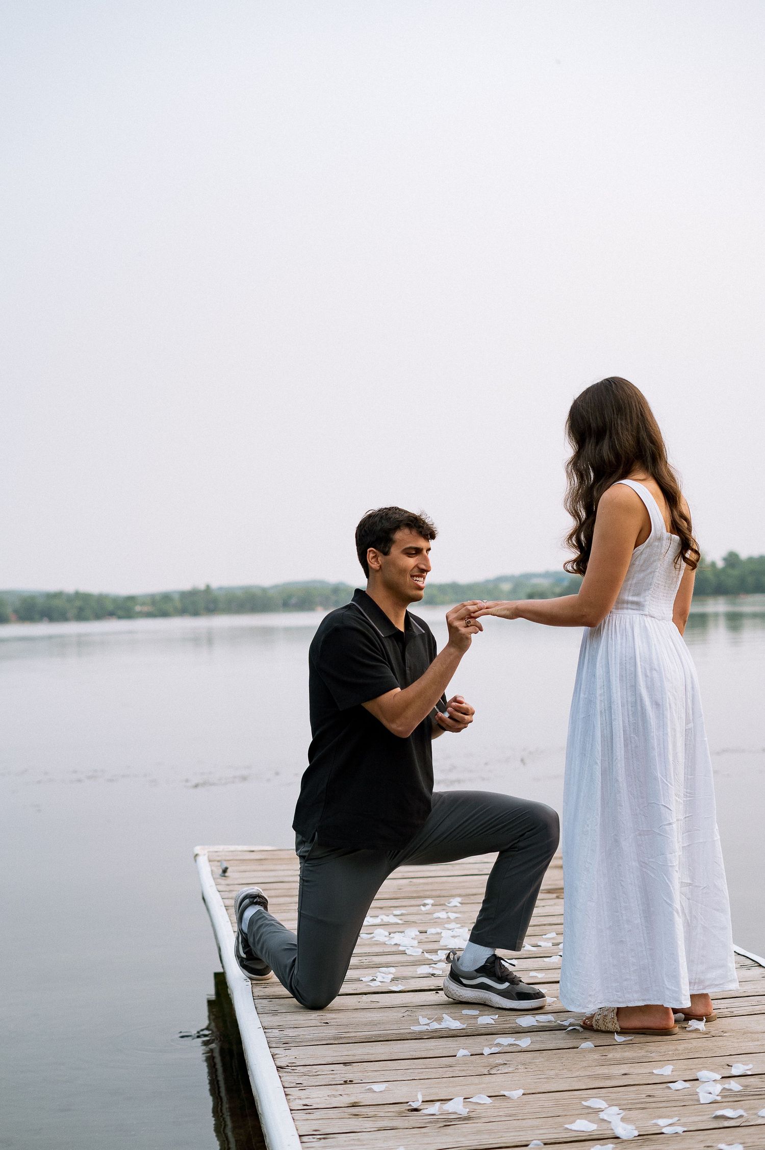 A romantic proposal sequence on a wooden dock by a lake, showing a person in black kneeling before someone in white.