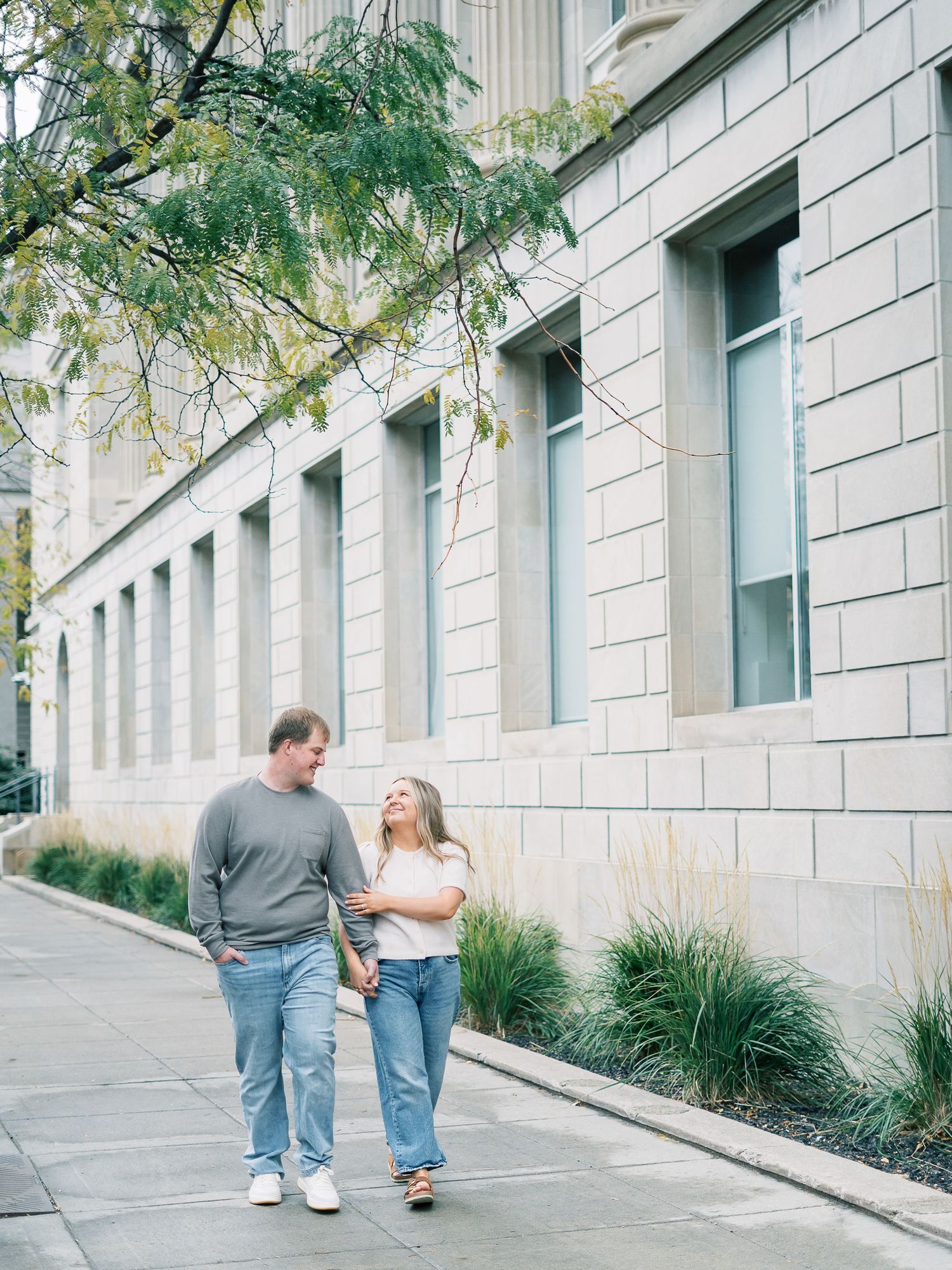 A couple walks together holding hands along a stone building with large windows and ornate architecture.
