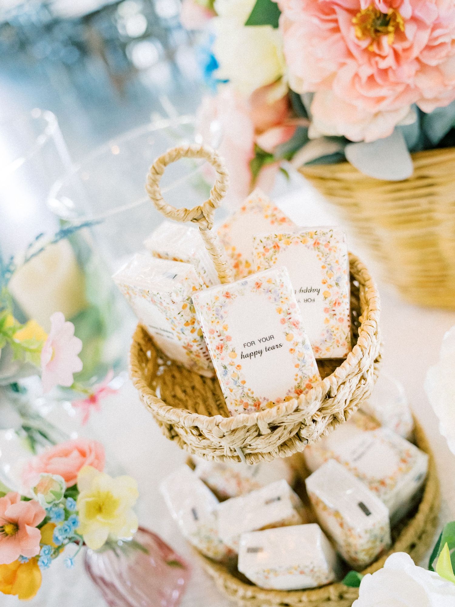 Tiered wicker basket display with floral wedding favors and seed packets surrounded by pink peonies and spring flowers.
