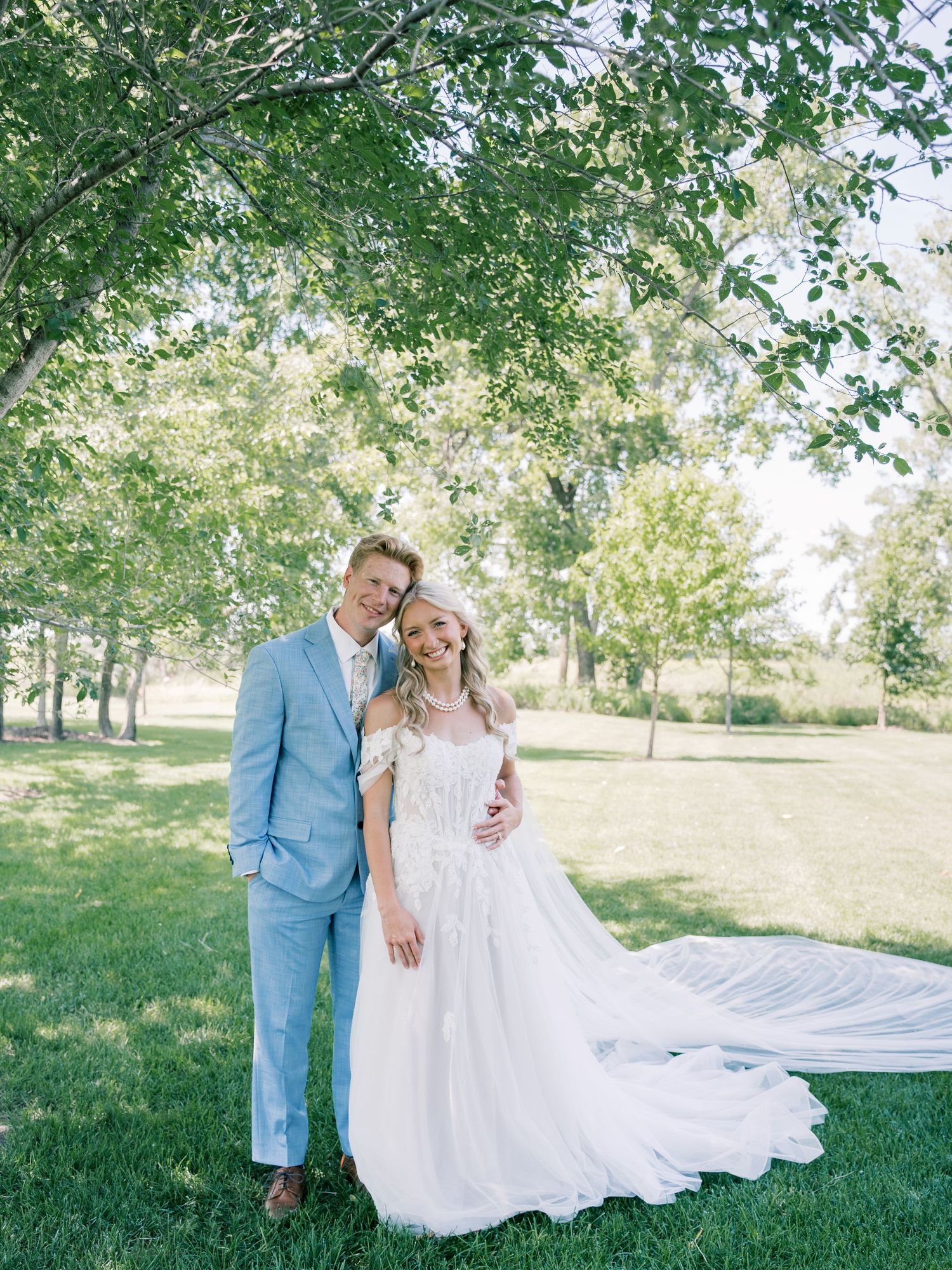 Bride in white off-shoulder gown with long train stands with groom in light blue suit outdoors under green trees.