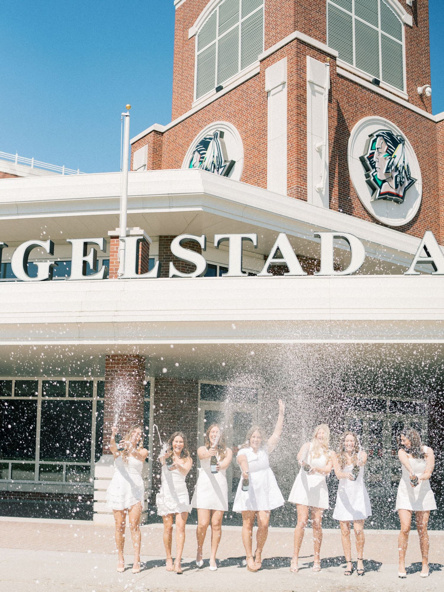 Women in white dresses popping champagne bottles outside Engelstad Arena, creating a festive celebration.