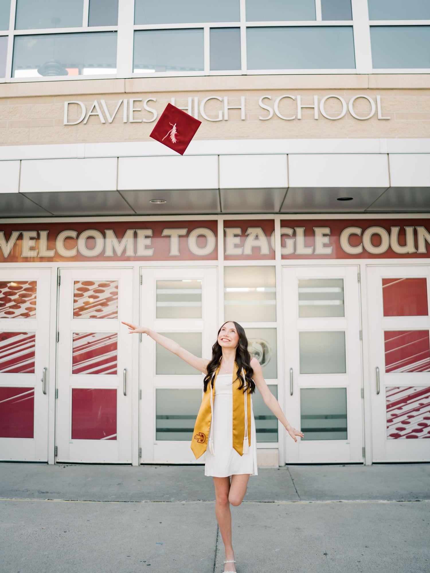 Graduate tosses cap in front of Davies High School Welcome to Eagle Country entrance in white dress and gold stole.
