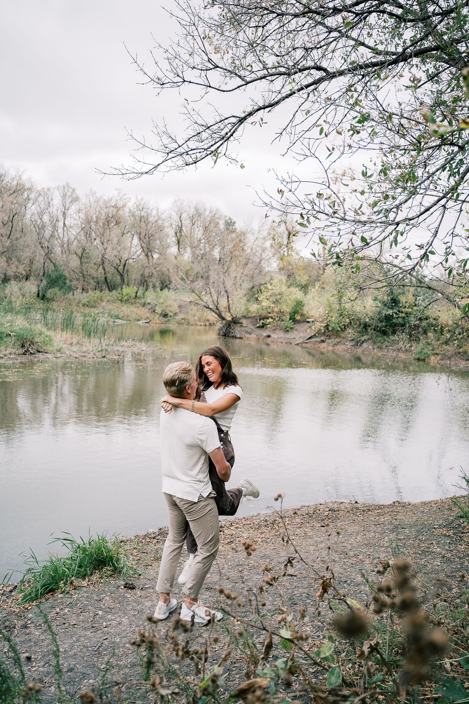A couple embraces on the rocky shore of a serene lake surrounded by bare winter trees.