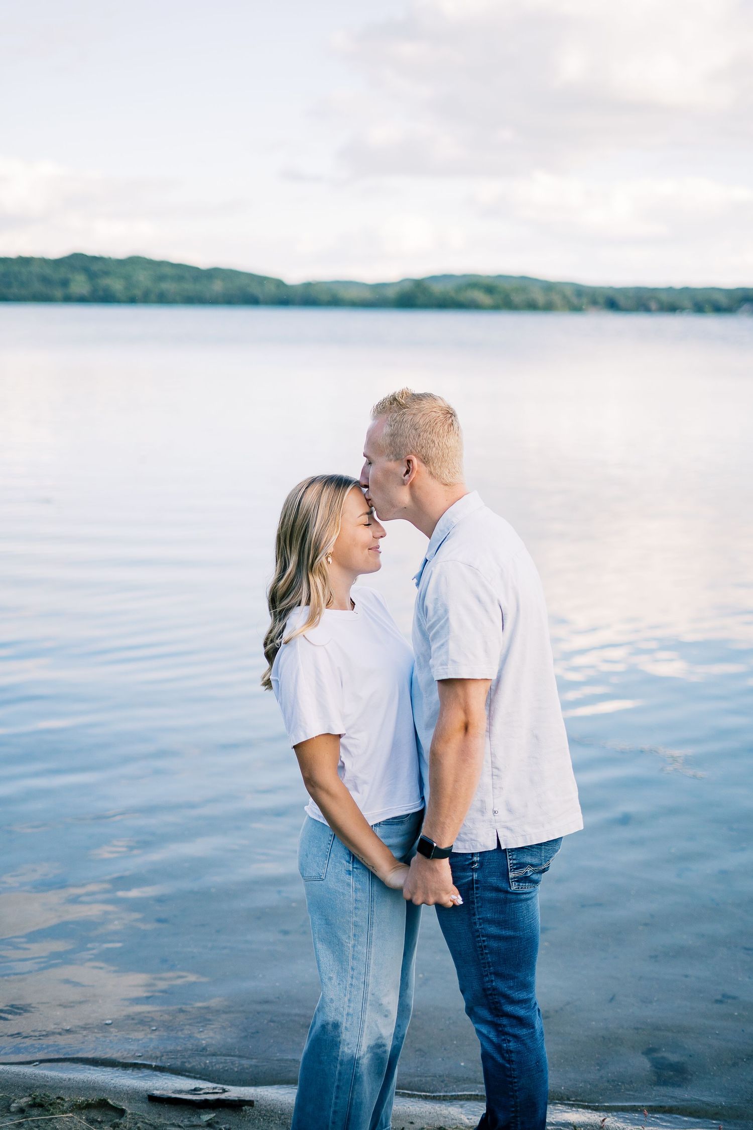 Romantic couple in white and denim share tender moments by a serene lakeside at sunset.