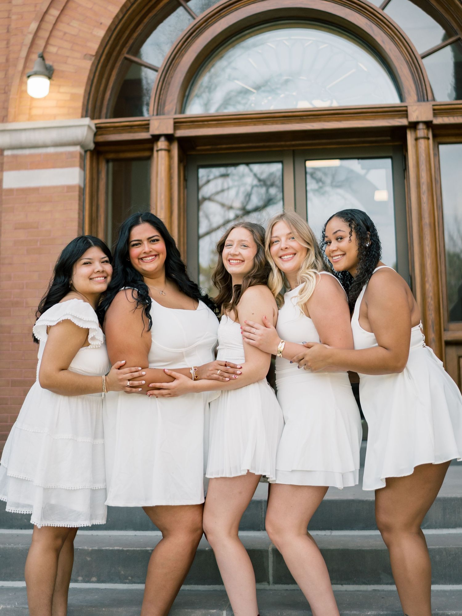 Five women in matching white mini dresses pose together smiling on steps outside a brick building.