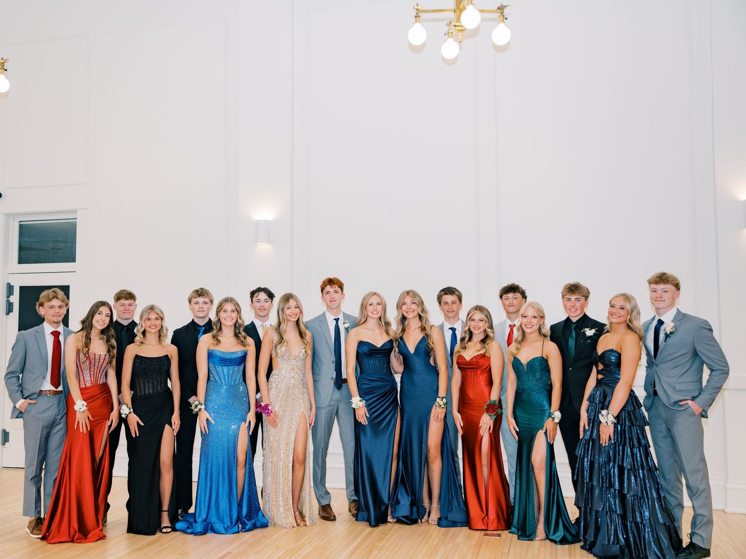 Group of teenagers in formal prom attire posing together in elegant white venue, wearing colorful gowns and suits.
