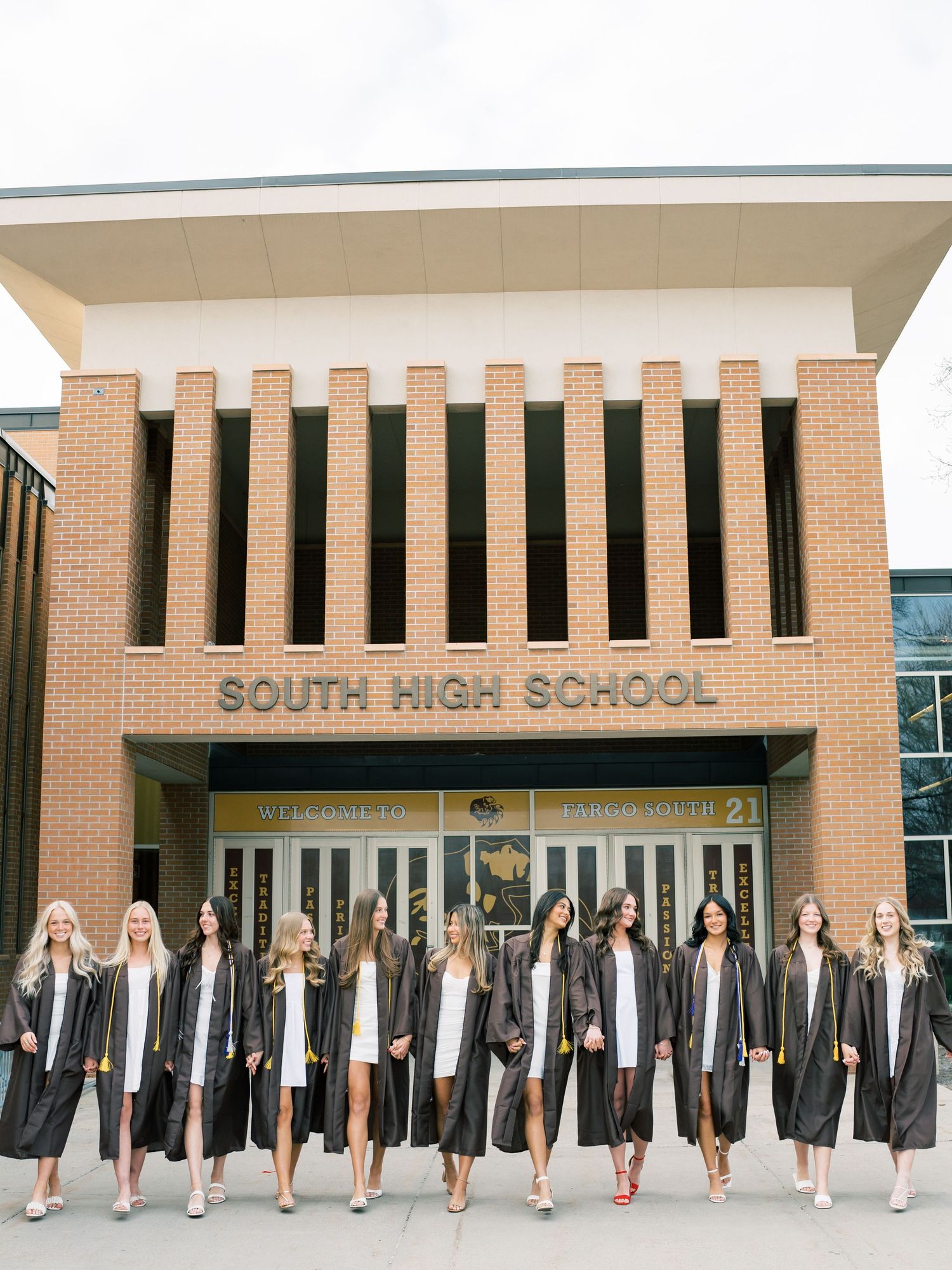Group of female graduates in black gowns posing in front of South High School building on graduation day.