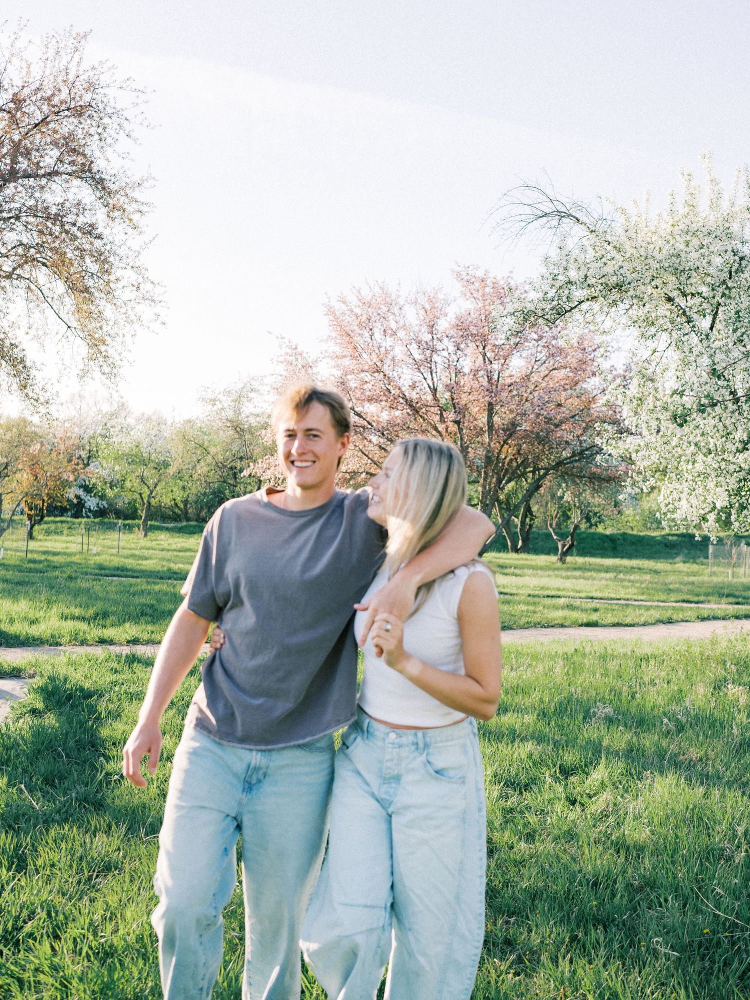 A smiling couple walks arm in arm through a lush green park with blooming trees on a sunny spring day.
