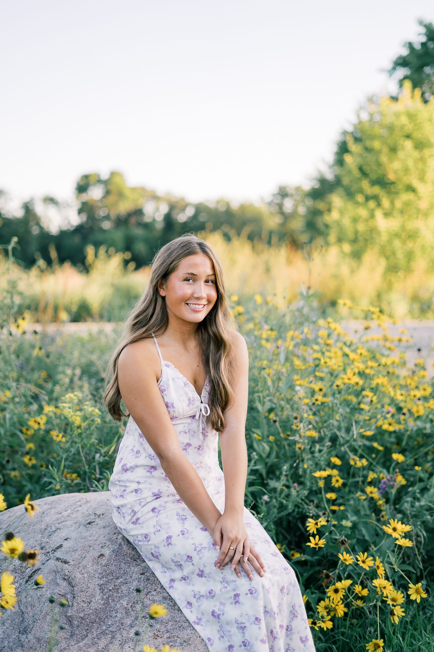 A person in a floral dress sits on a large rock surrounded by blooming yellow wildflowers in a sunlit meadow.