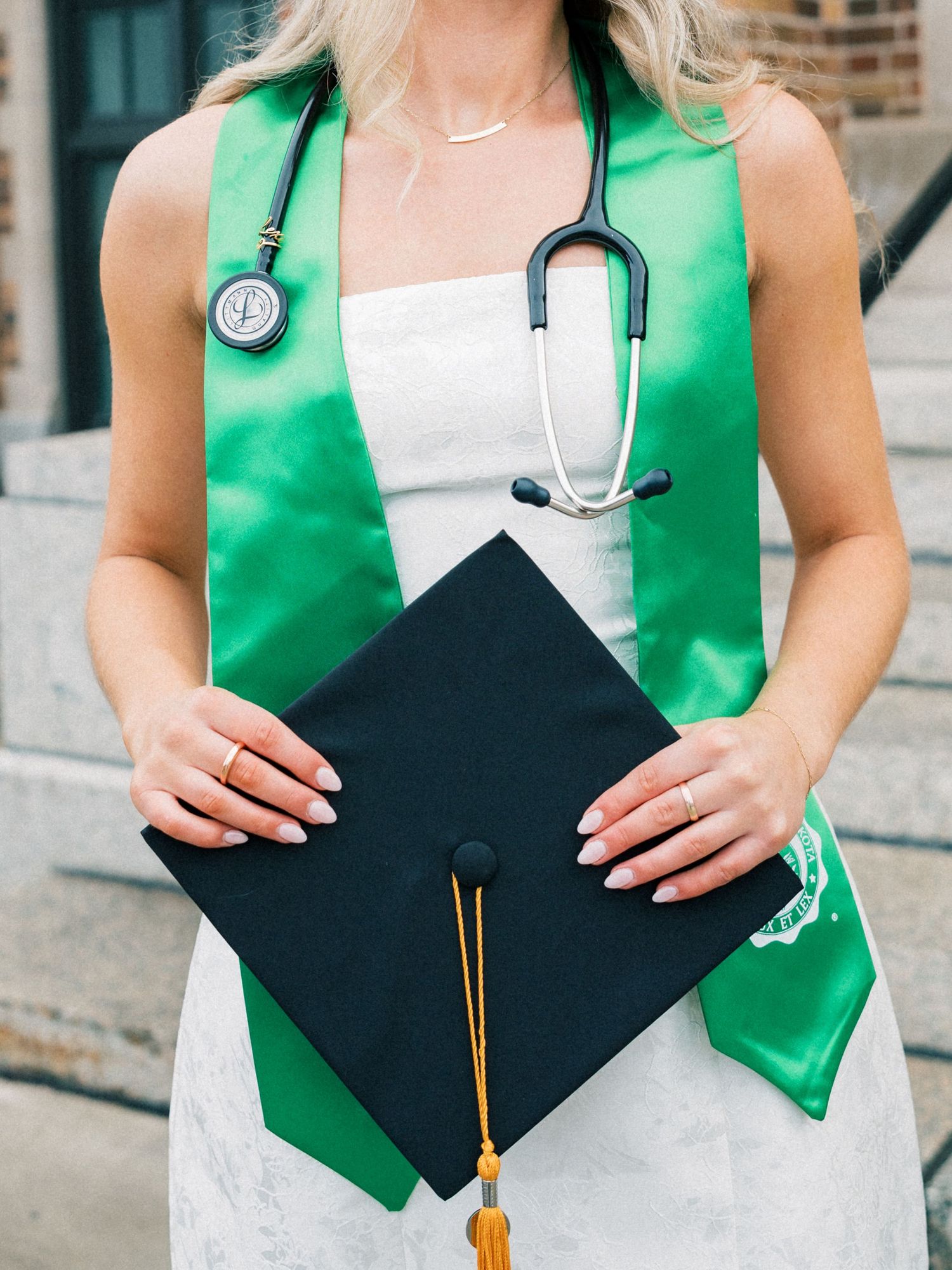 Graduate holding black cap with gold tassel, wearing green sash and stethoscope over white dress.