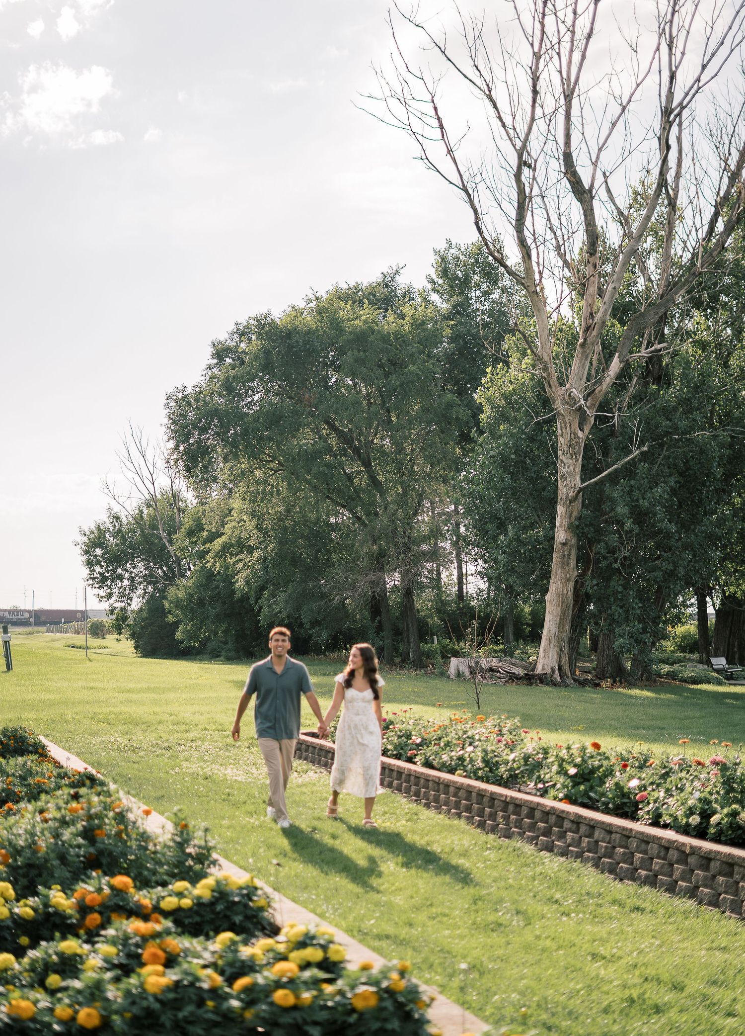 A couple walks together on a lush green lawn bordered by vibrant marigold flower beds and tall trees on a summer day.