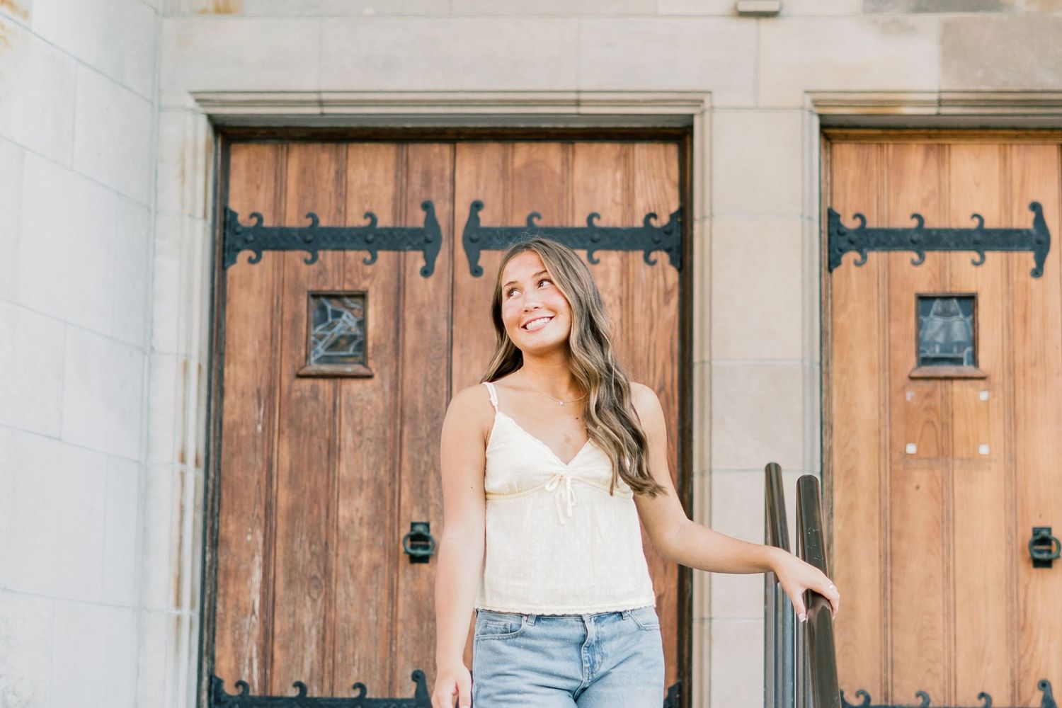 Smiling person in white top and jeans stands against rustic wooden doors with decorative iron hinges.