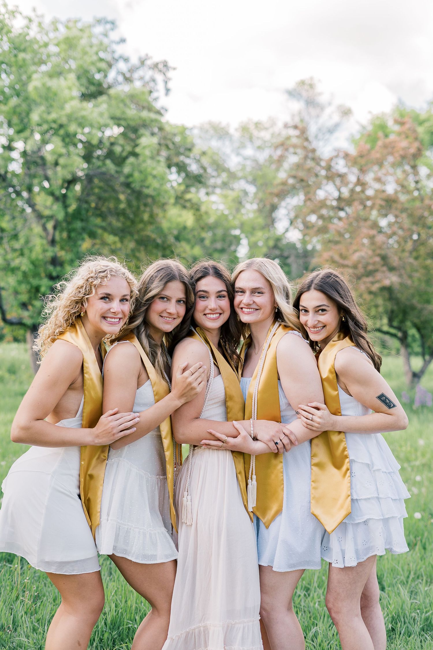 A senior portrait group wearing white dresses and gold sashes pose together outdoors in a natural garden setting.