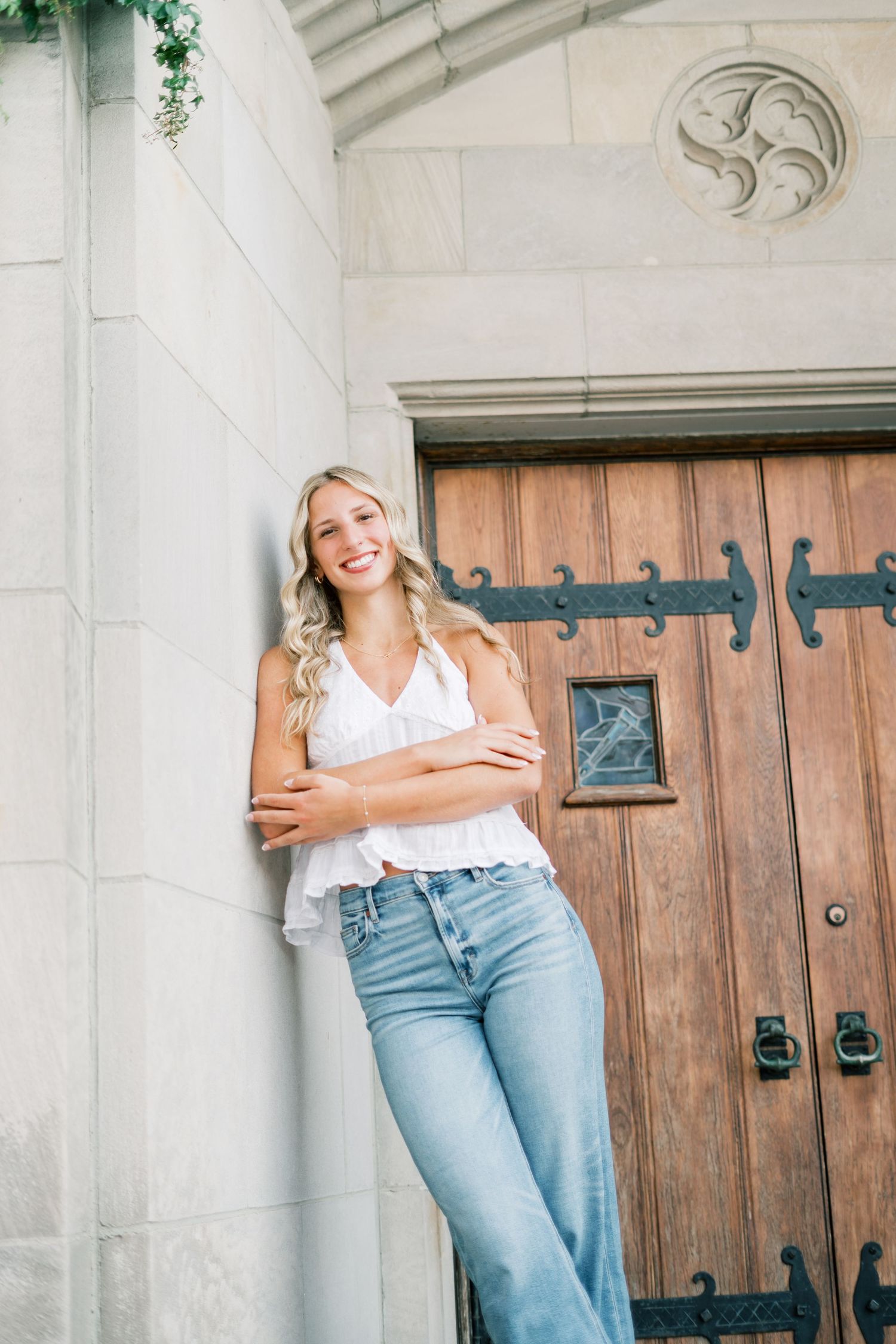 Woman in white top and light blue jeans poses against stone wall with wooden door in background.