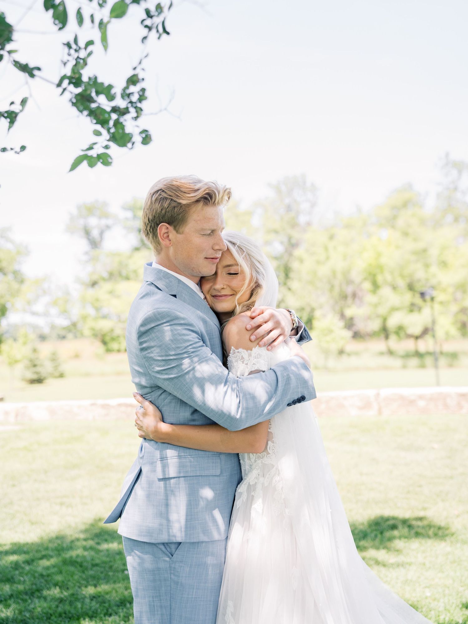 A bride and groom share a tender embrace outdoors on their wedding day, surrounded by lush green trees in soft natural light.