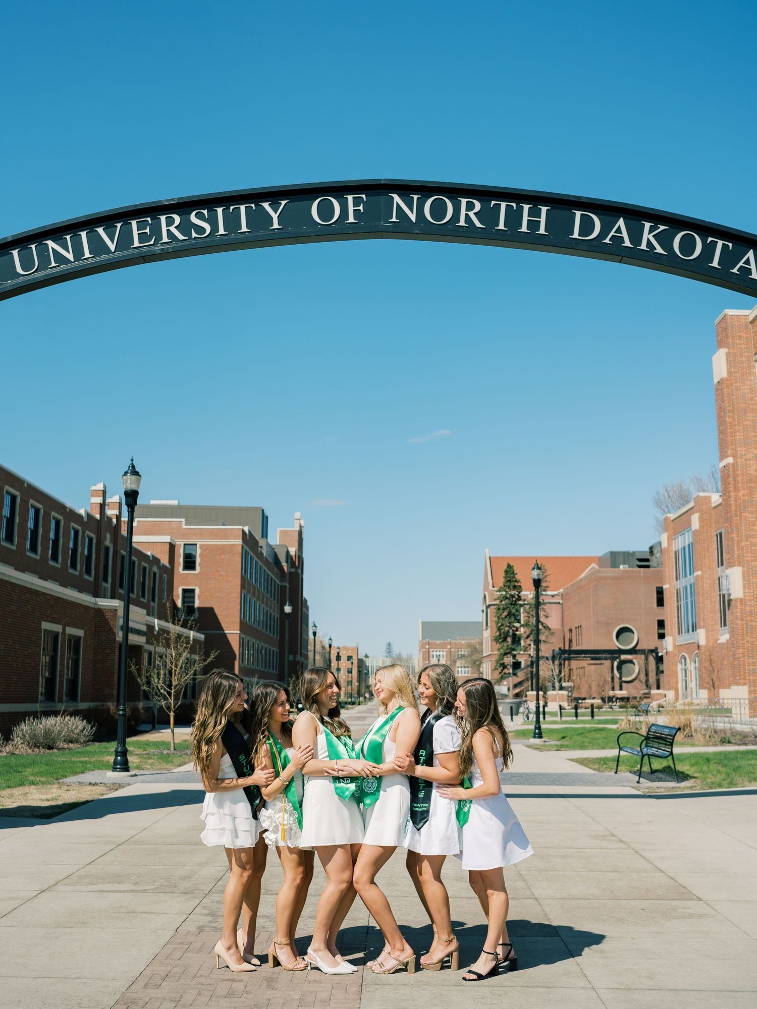 Group of women in green and white outfits posing under University of North Dakota arch on a sunny day.