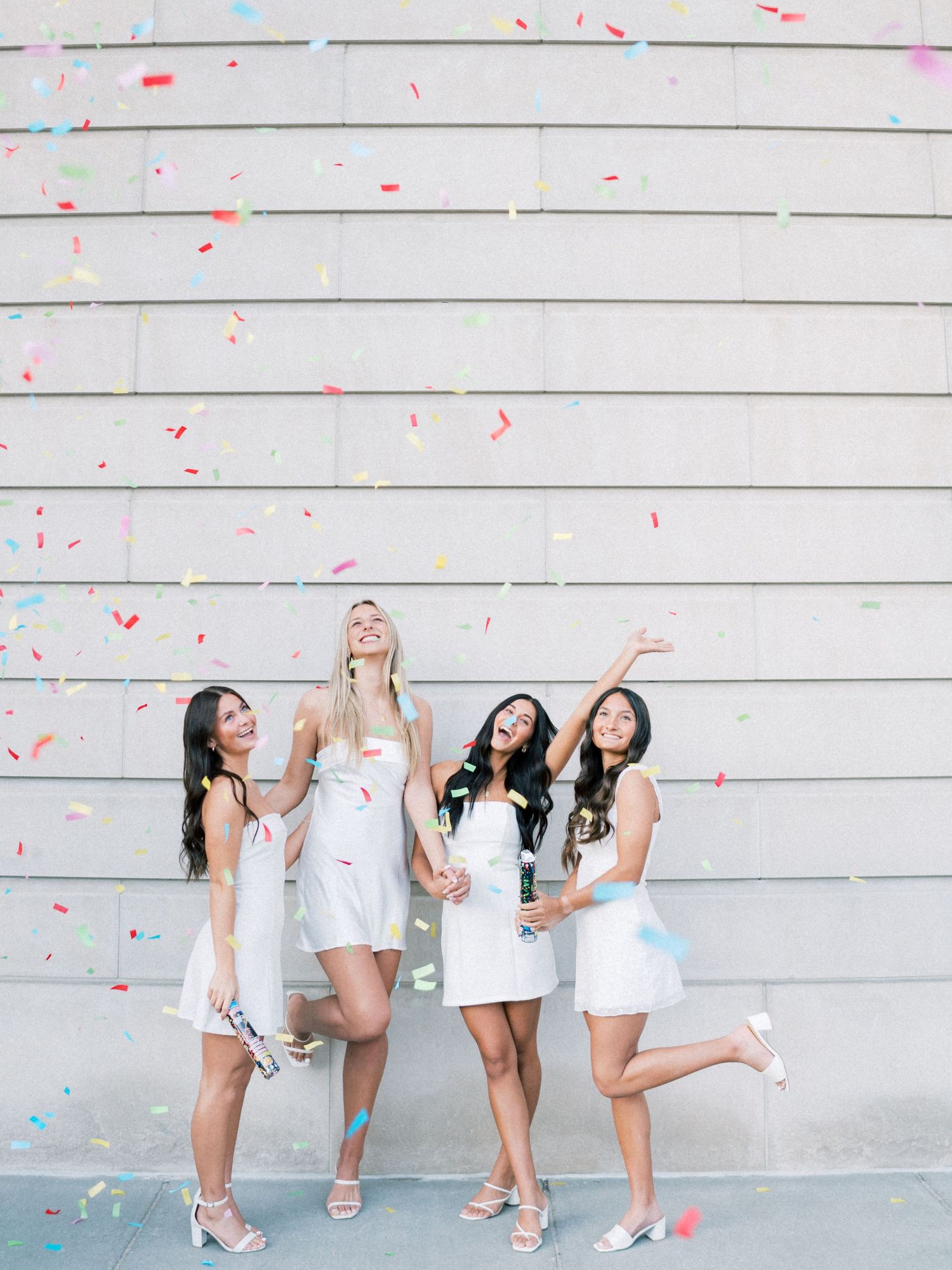 Four women in white dresses celebrate with colorful confetti falling around them against a gray wall.