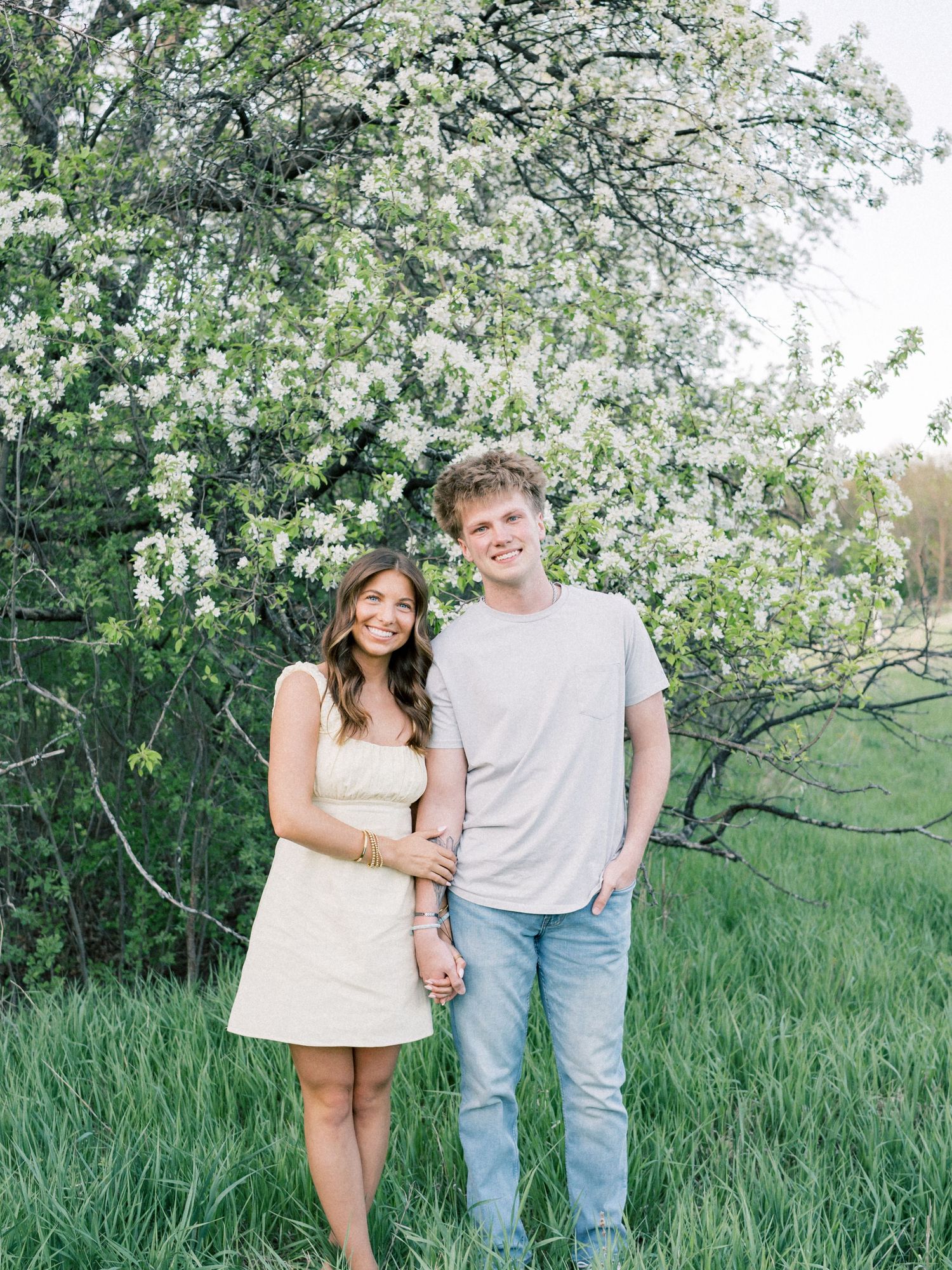 A smiling couple poses in a green field beside a blooming white flowering tree on a bright spring day.