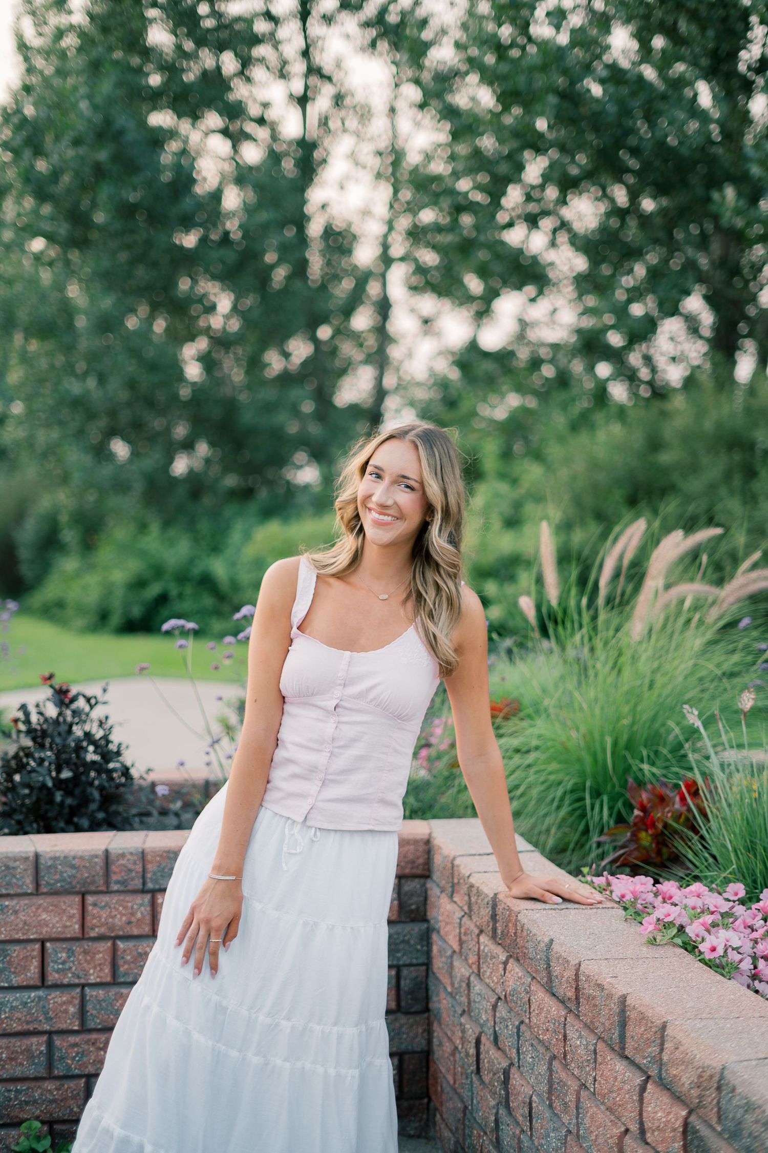 A person in a white summer dress poses gracefully against a brick retaining wall with ornamental grasses and flowers.