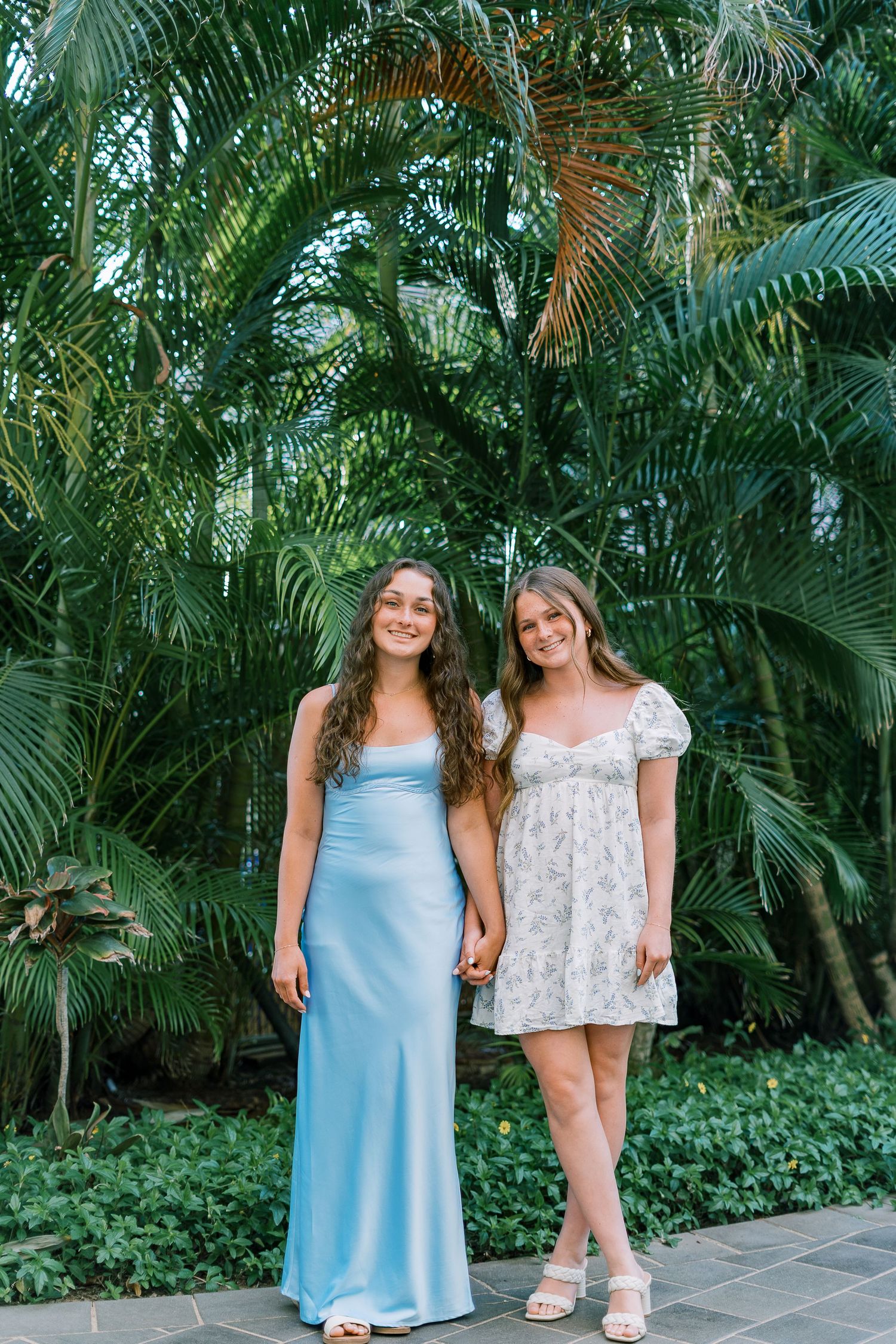 Two companions stand together in front of a tropical palm garden backdrop.