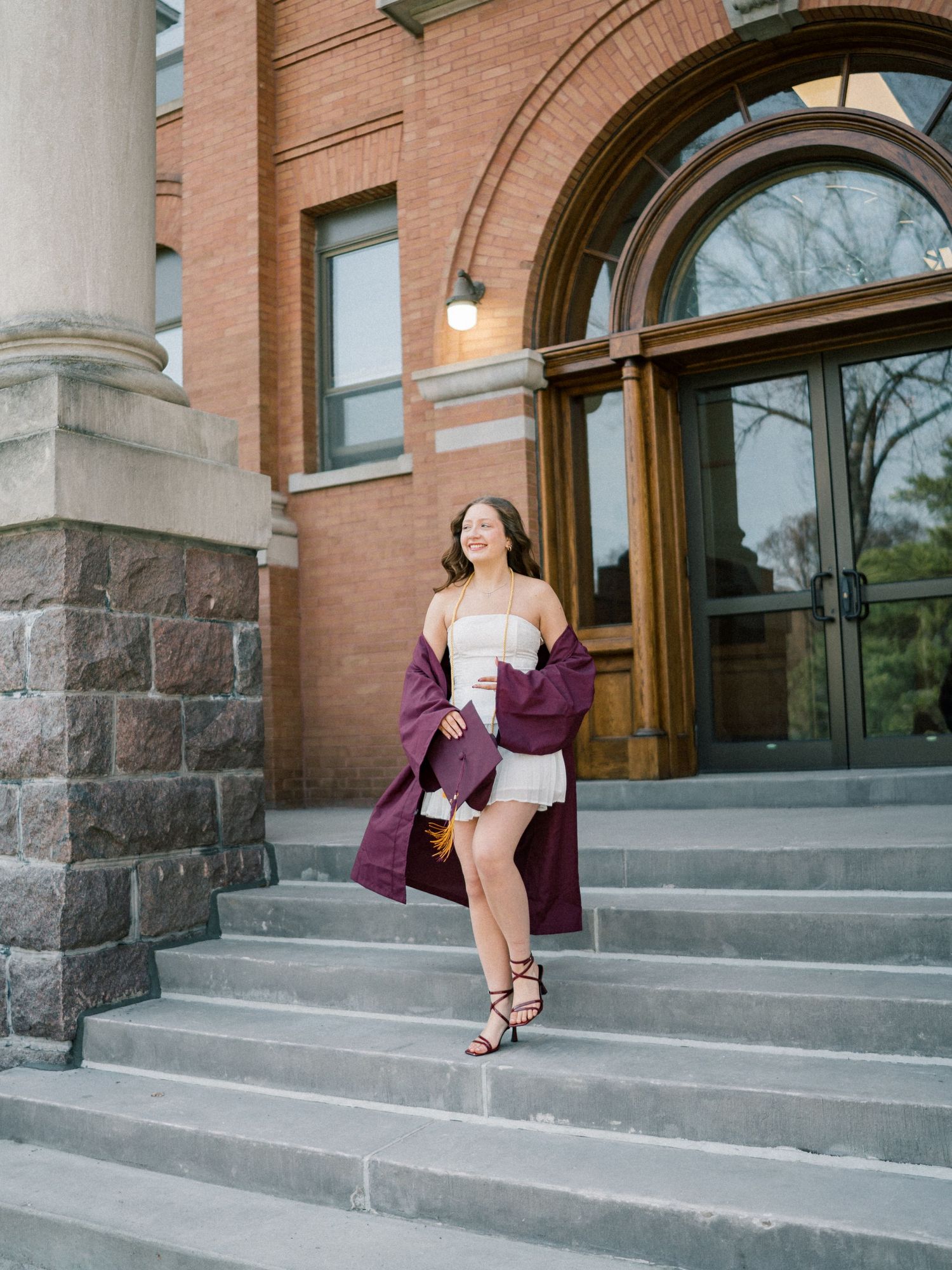 Smiling graduate in white dress and burgundy gown walks down steps of brick university building entrance.