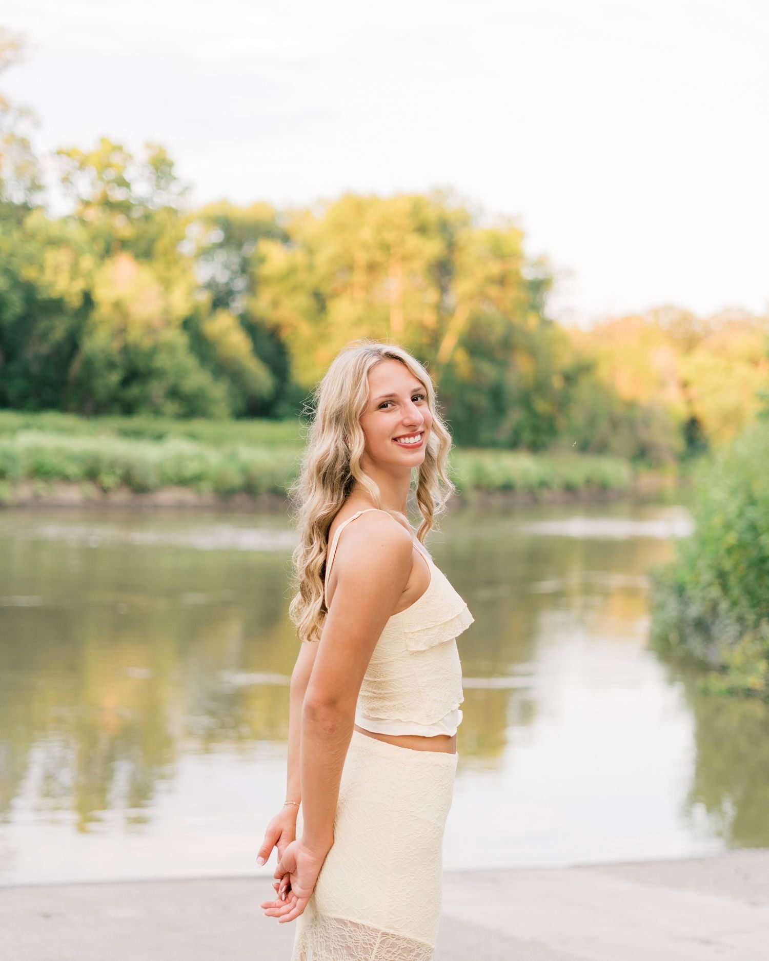 A person in cream-colored attire poses joyfully by a serene lake during golden hour with trees in the background.