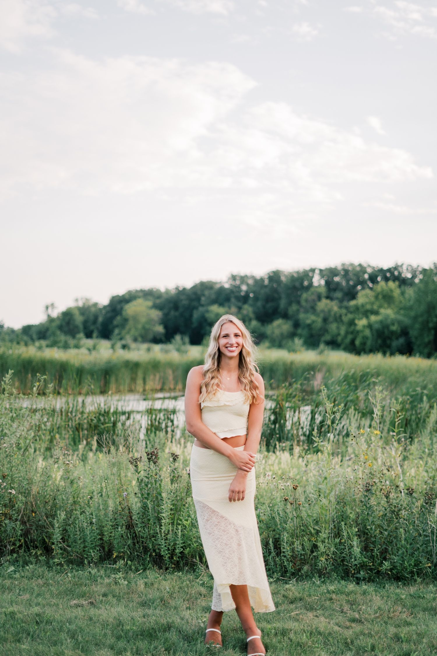 A woman in a white dress poses in a scenic meadow filled with wildflowers and tall grass during golden hour.