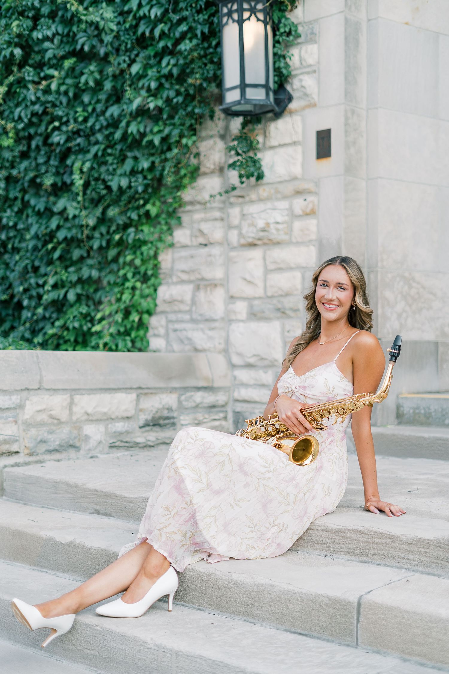 A musician in a white lace dress poses with a saxophone on stone steps with ivy-covered walls.