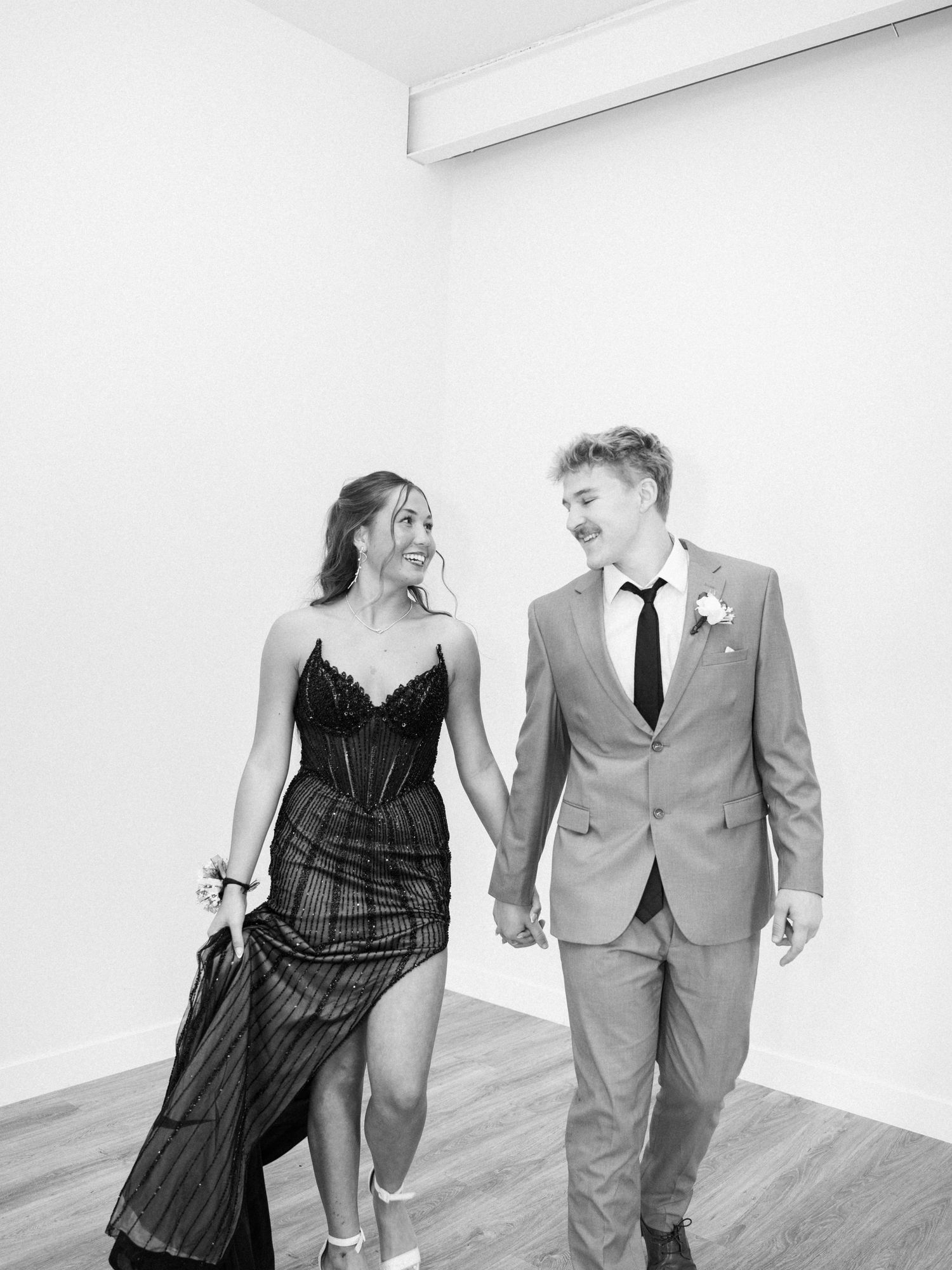 Black and white photo of a smiling couple in formal wear holding hands and walking together at prom.