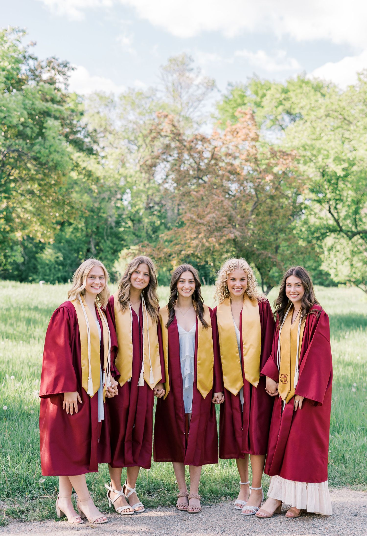 Graduates in burgundy graduation gowns and gold stoles pose together outdoors on a sunny spring day.