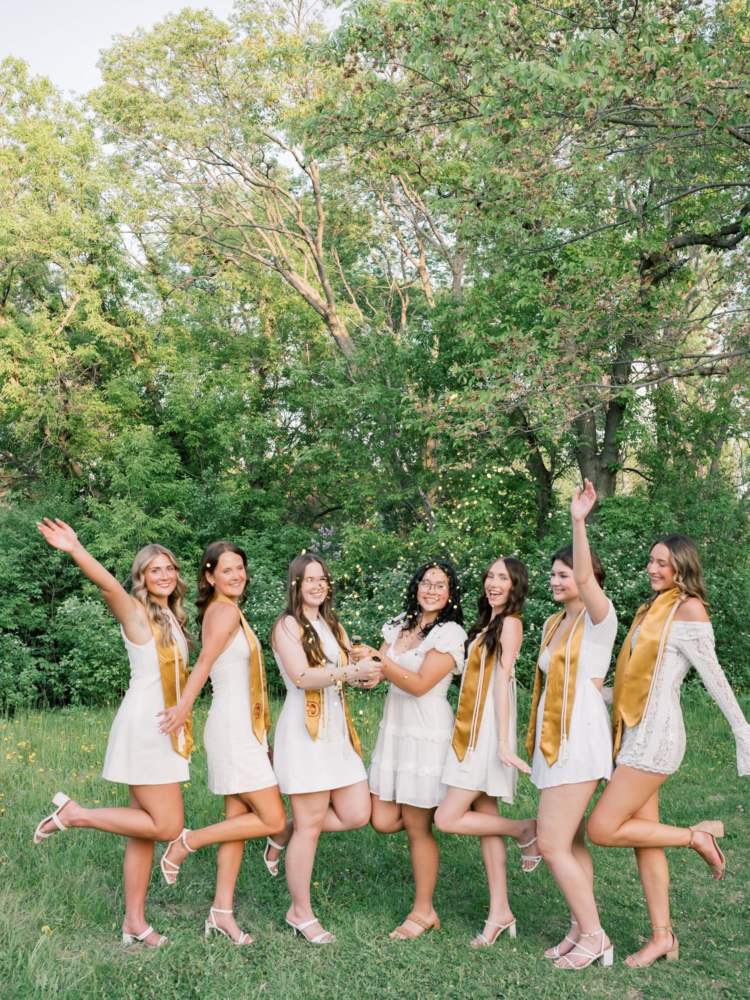 Seven women in white dresses and gold graduation stoles celebrate outdoors in a lush green setting.