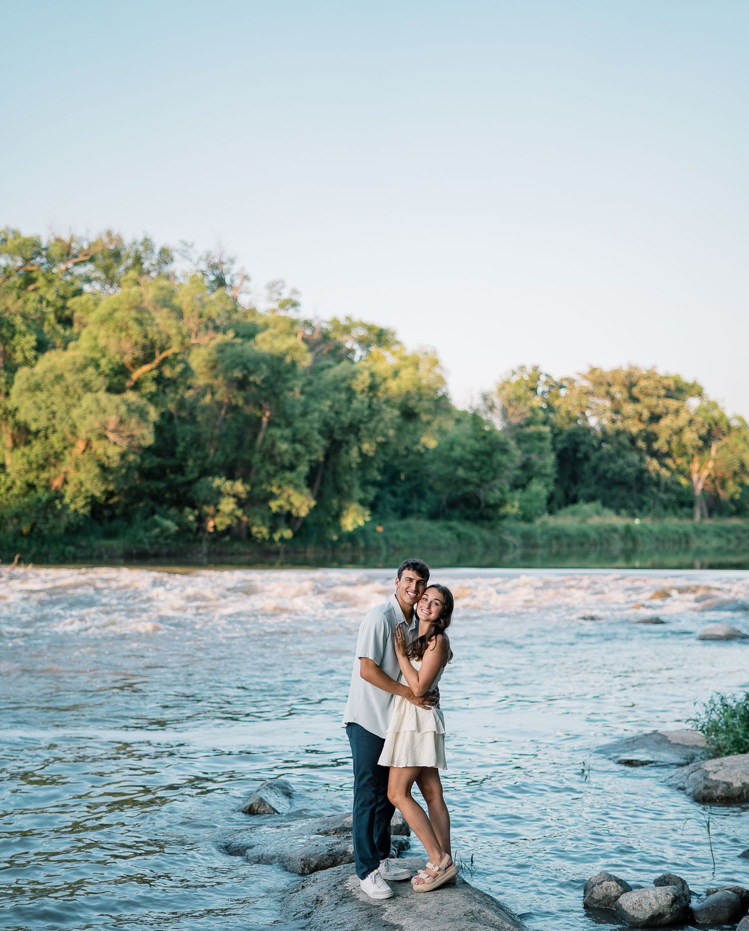 Couple shares romantic moments by a scenic river with trees and rocks during sunset.
