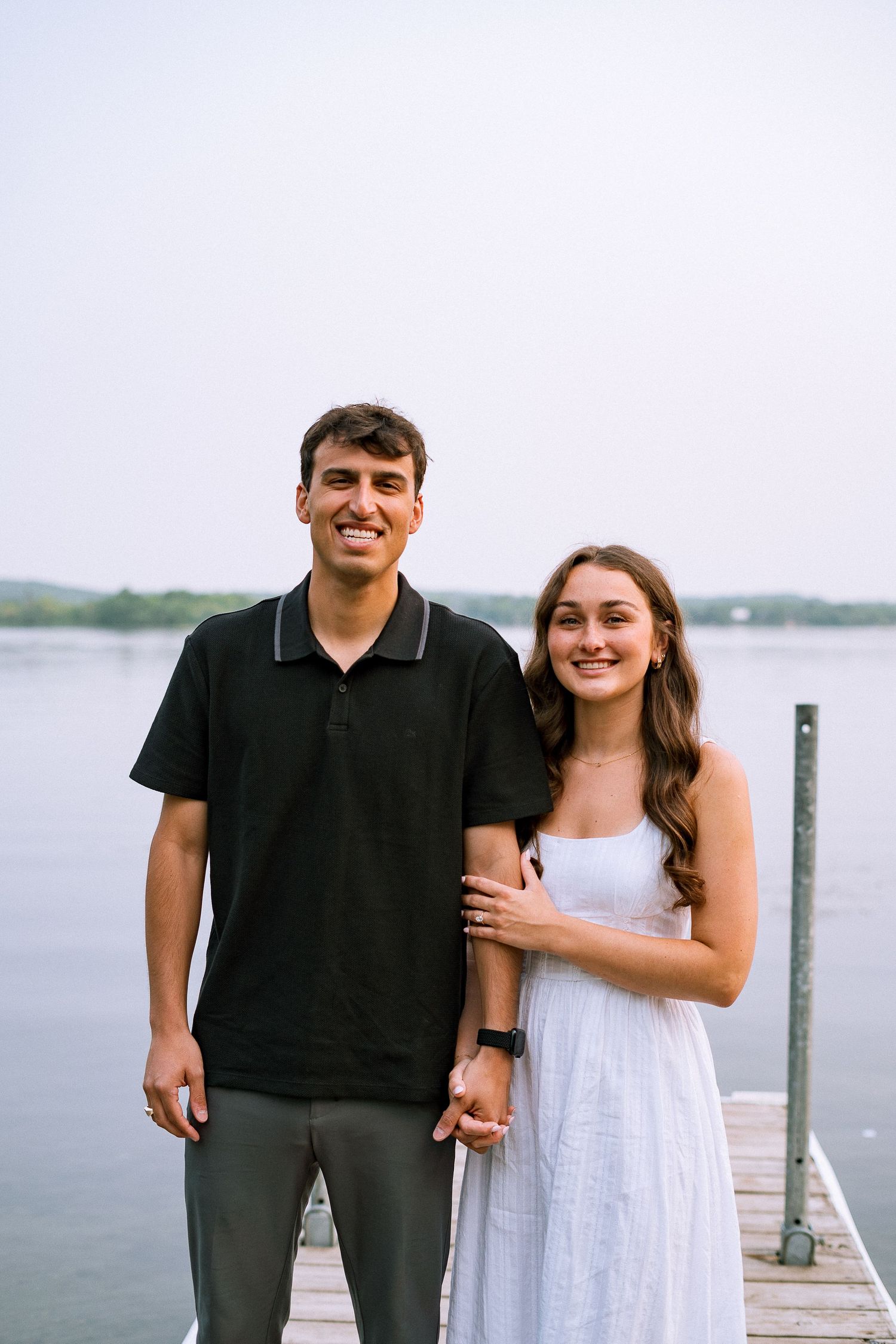 A couple poses together on a wooden dock by a lake, with the person in black and person in white smiling.