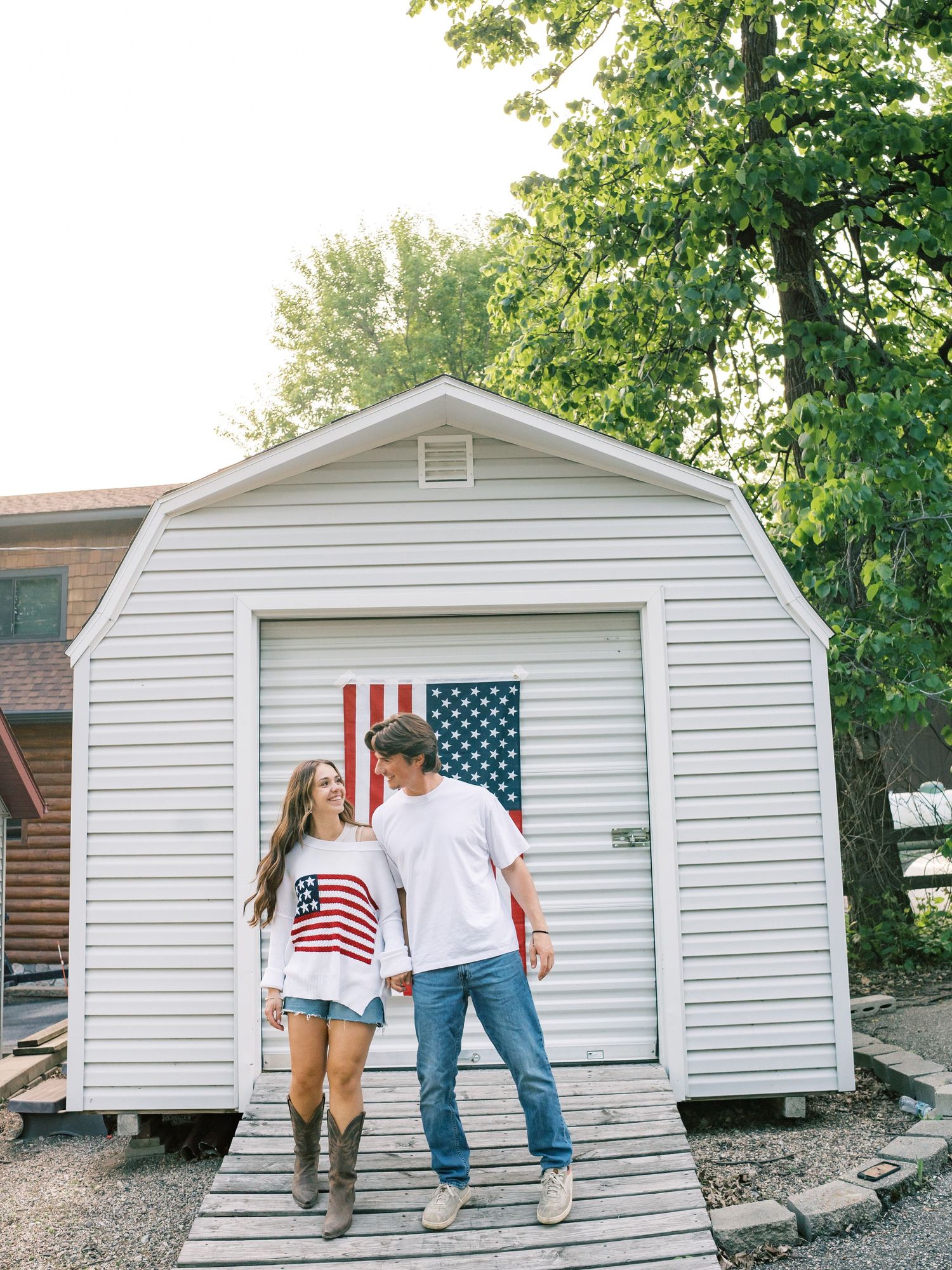 Couple in matching American flag outfits standing on wooden deck in front of white shed with US flag door.