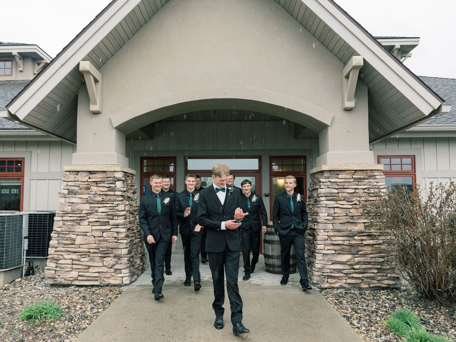 Groomsmen in black tuxedos with green boutonnieres exit a stone and stucco venue during a wedding.
