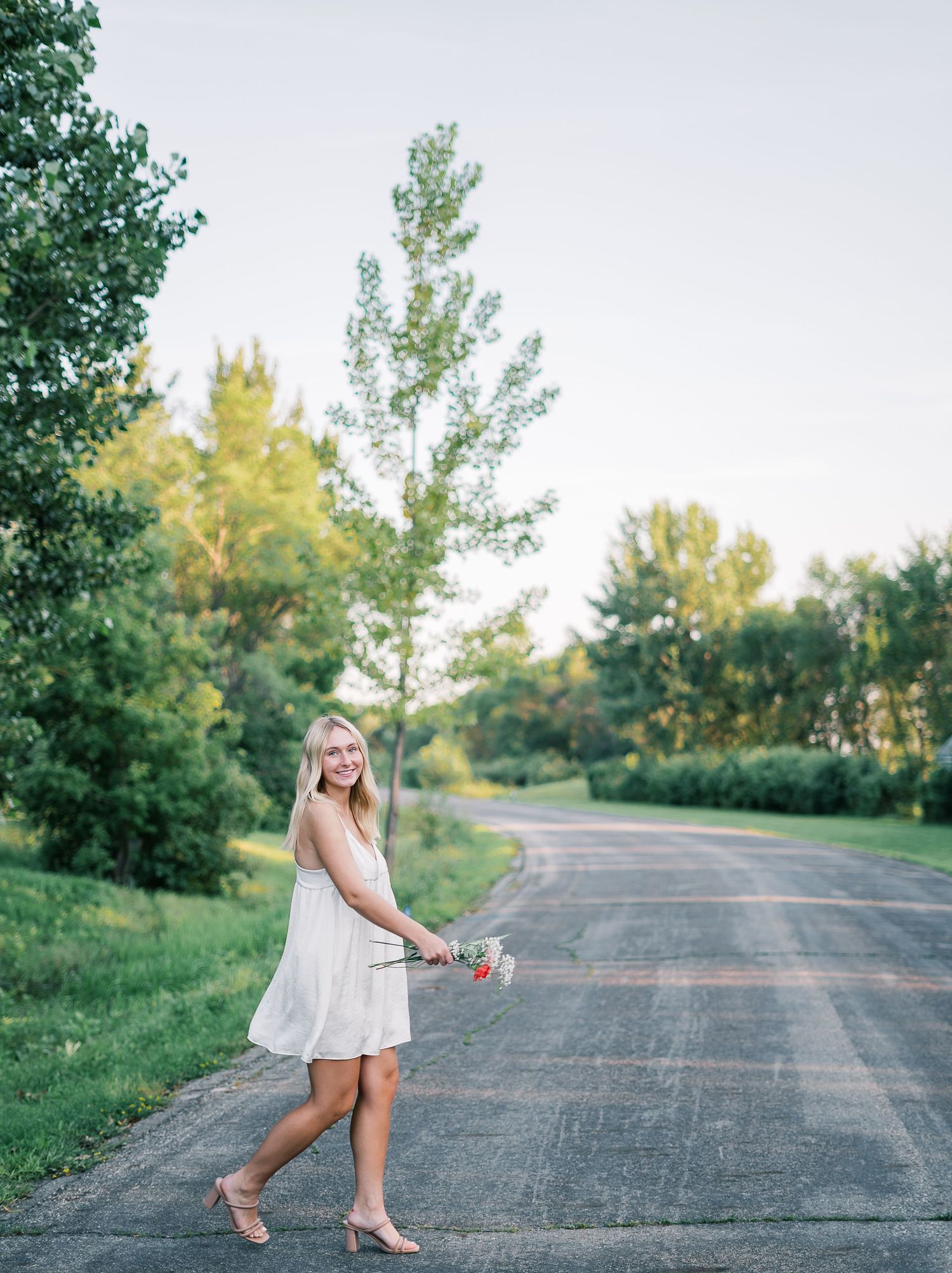 A person in a white dress walks along a winding country road surrounded by green trees on a summer day.