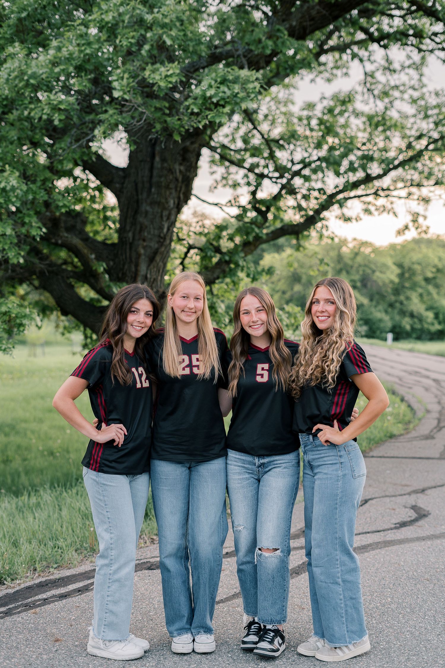 Four teammates in numbered black jerseys and jeans smile while posing on a winding path under trees.
