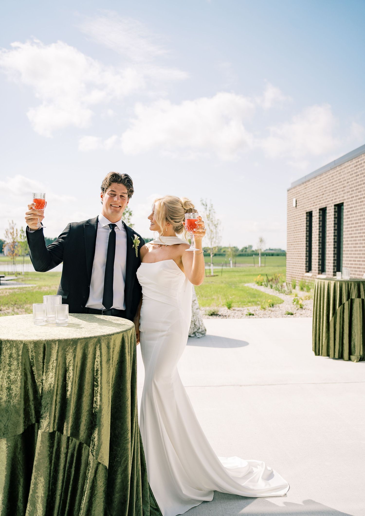 Bride and groom share a celebratory toast in front of rustic wooden fence outside a white brick building on sunny day.