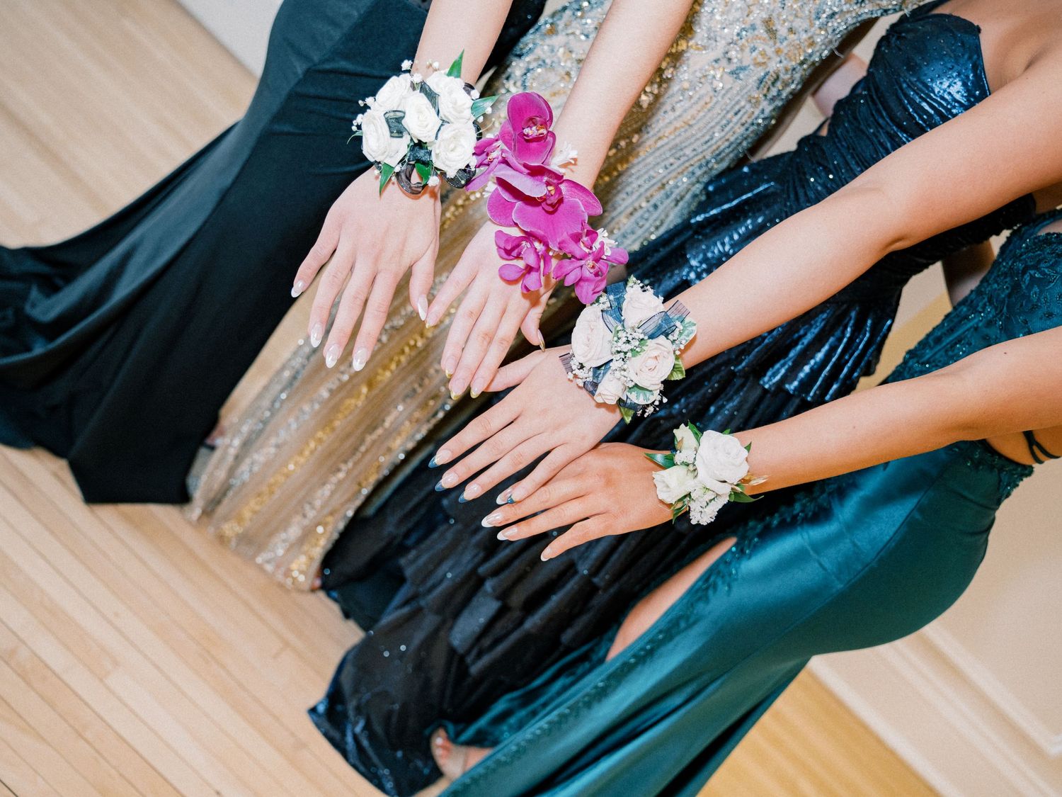 Four women in formal gowns display floral wrist corsages in pink and white blooms at prom.