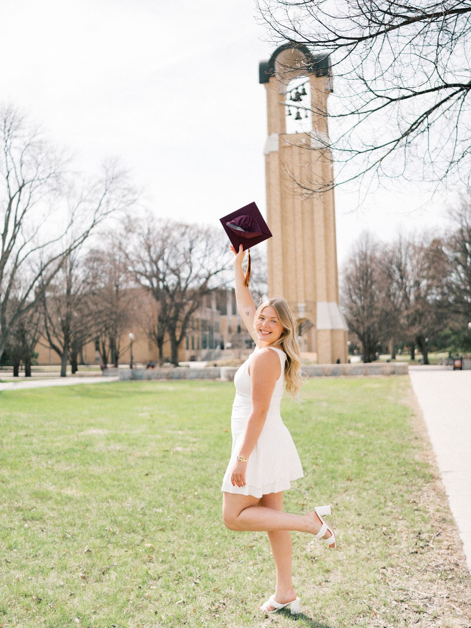 Smiling graduate in white dress tosses maroon cap in air near university bell tower on campus.