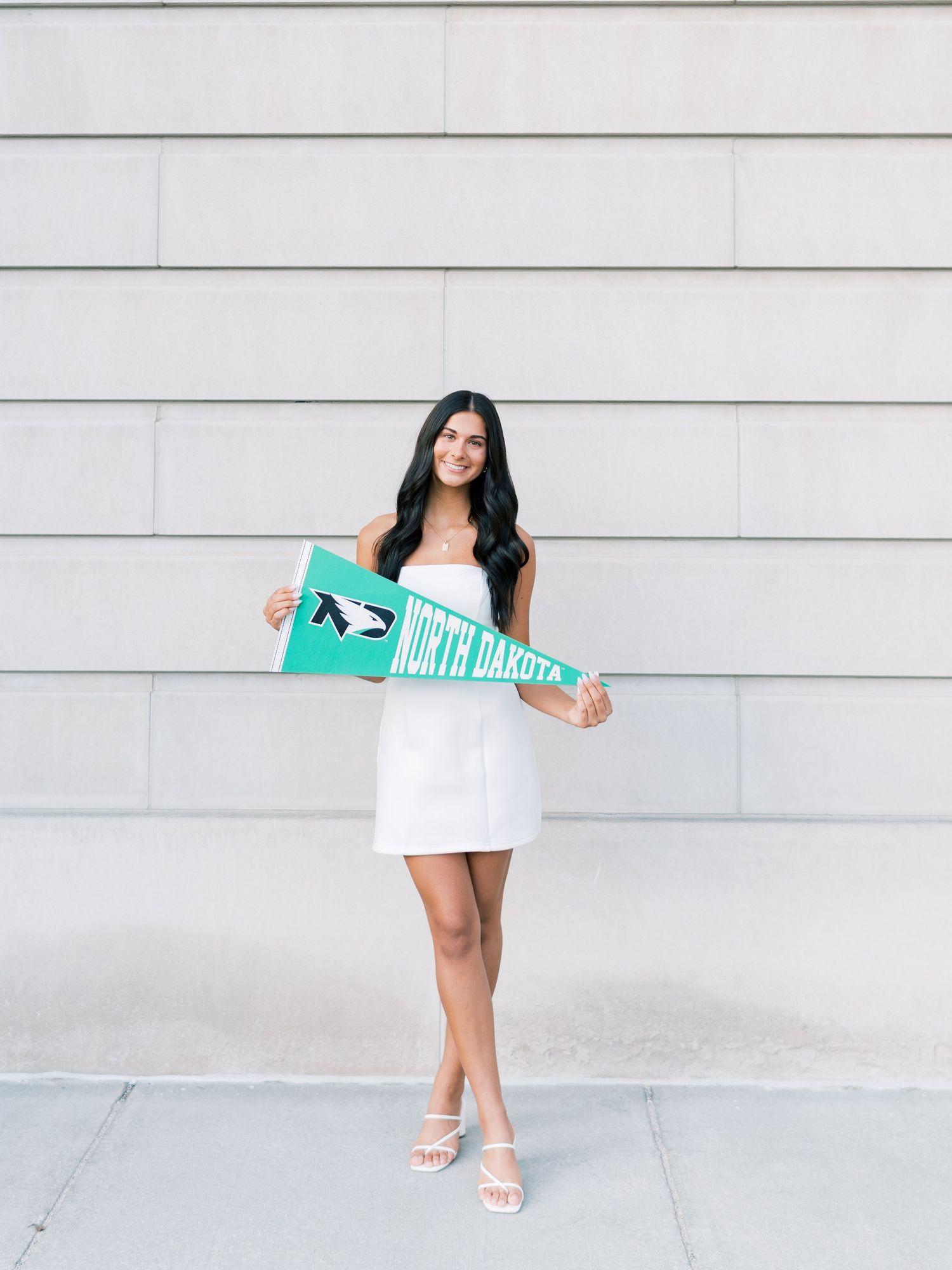 Young woman in white dress holding a green North Dakota university pennant flag, posing outdoors.