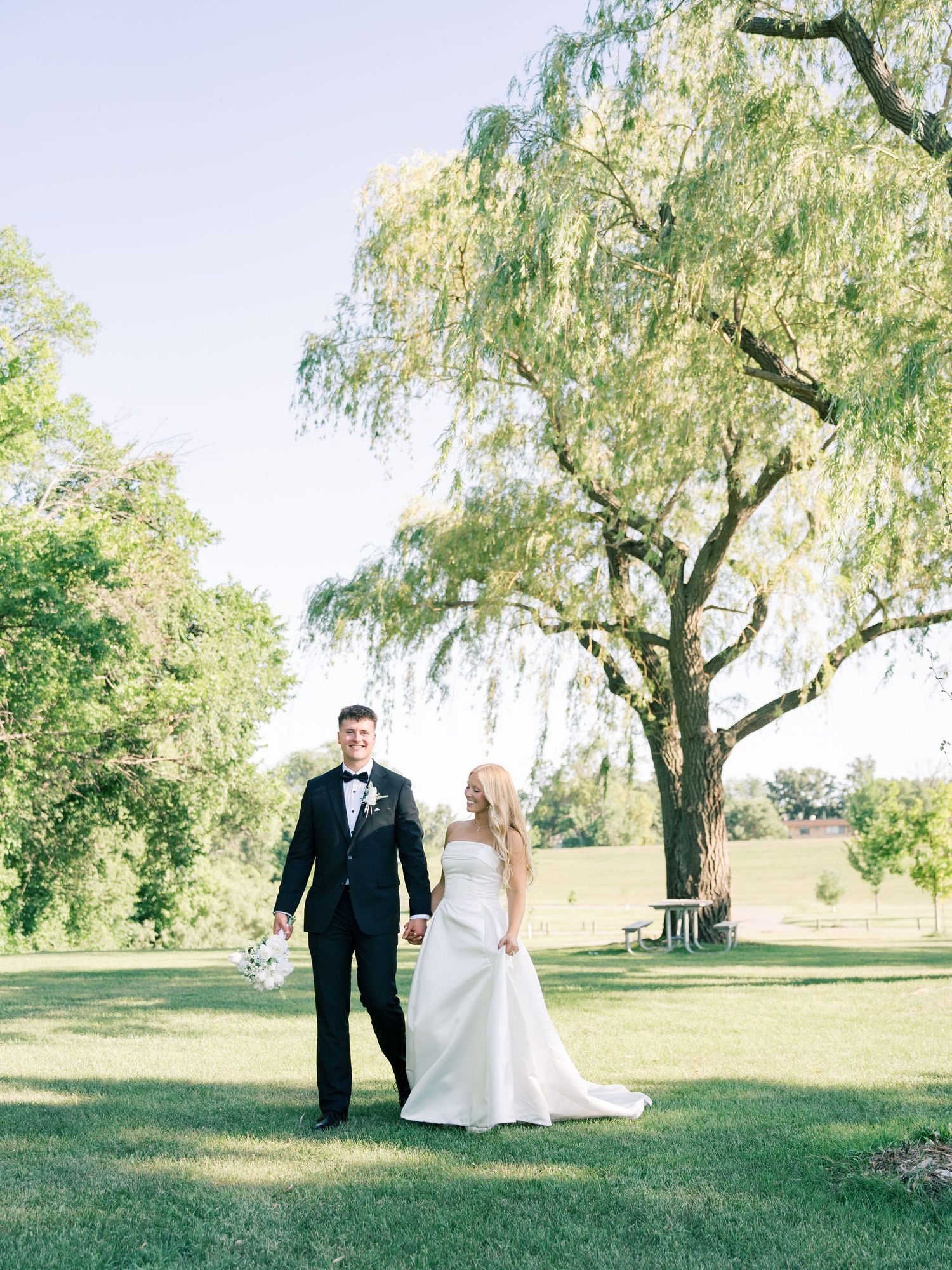 A bride and groom hold hands walking on green grass beneath a weeping willow tree on their sunny wedding day.