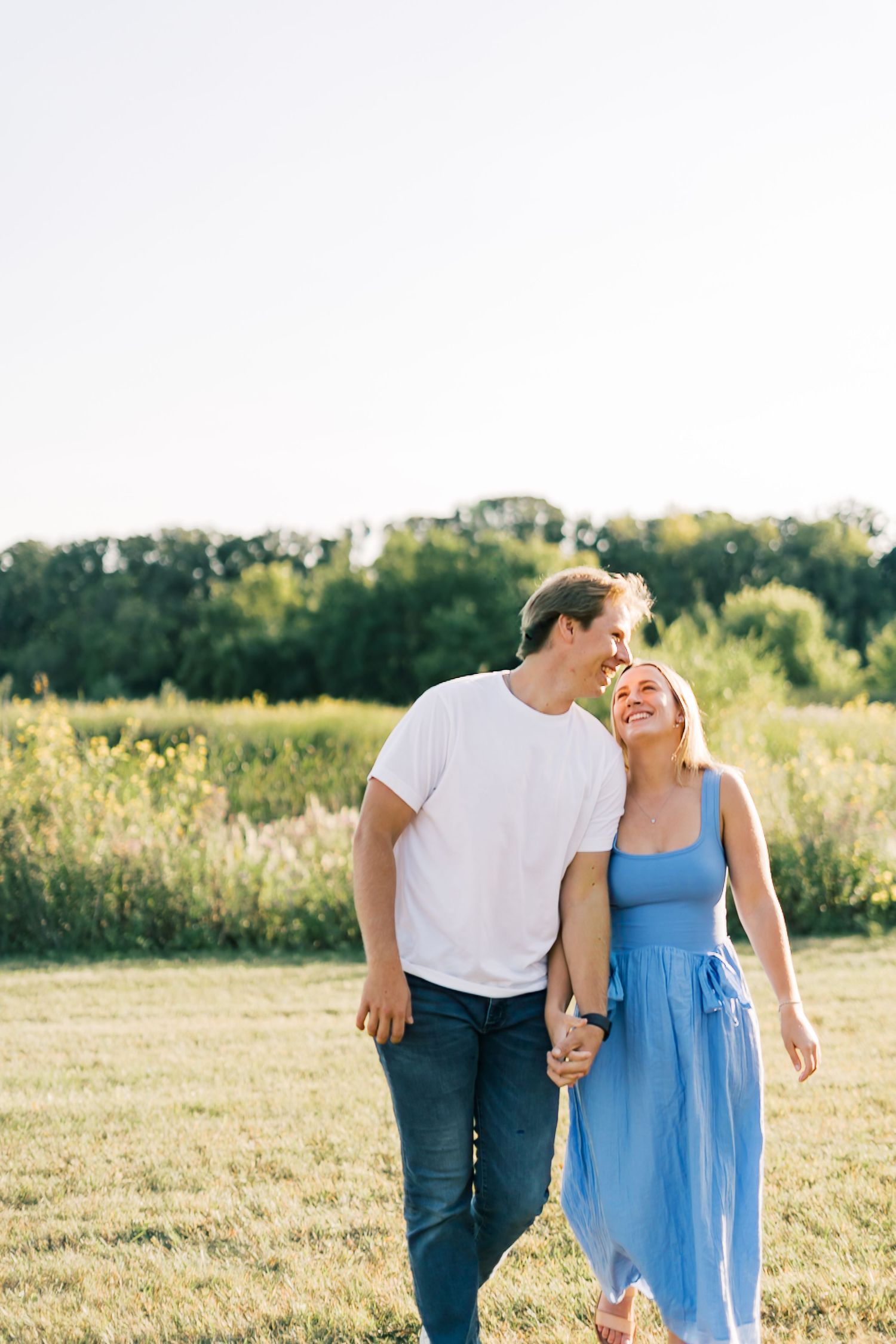 A couple walks together in a sunny meadow, the woman wearing a blue dress and the man in a white shirt.