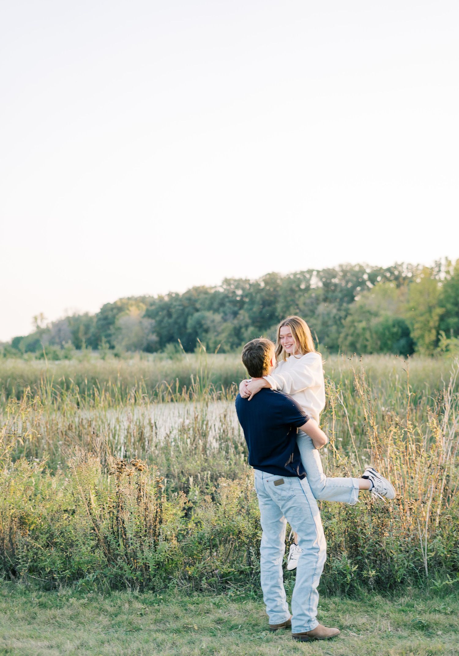 A couple shares a tender moment in a sunlit meadow with tall grass and trees in the background.