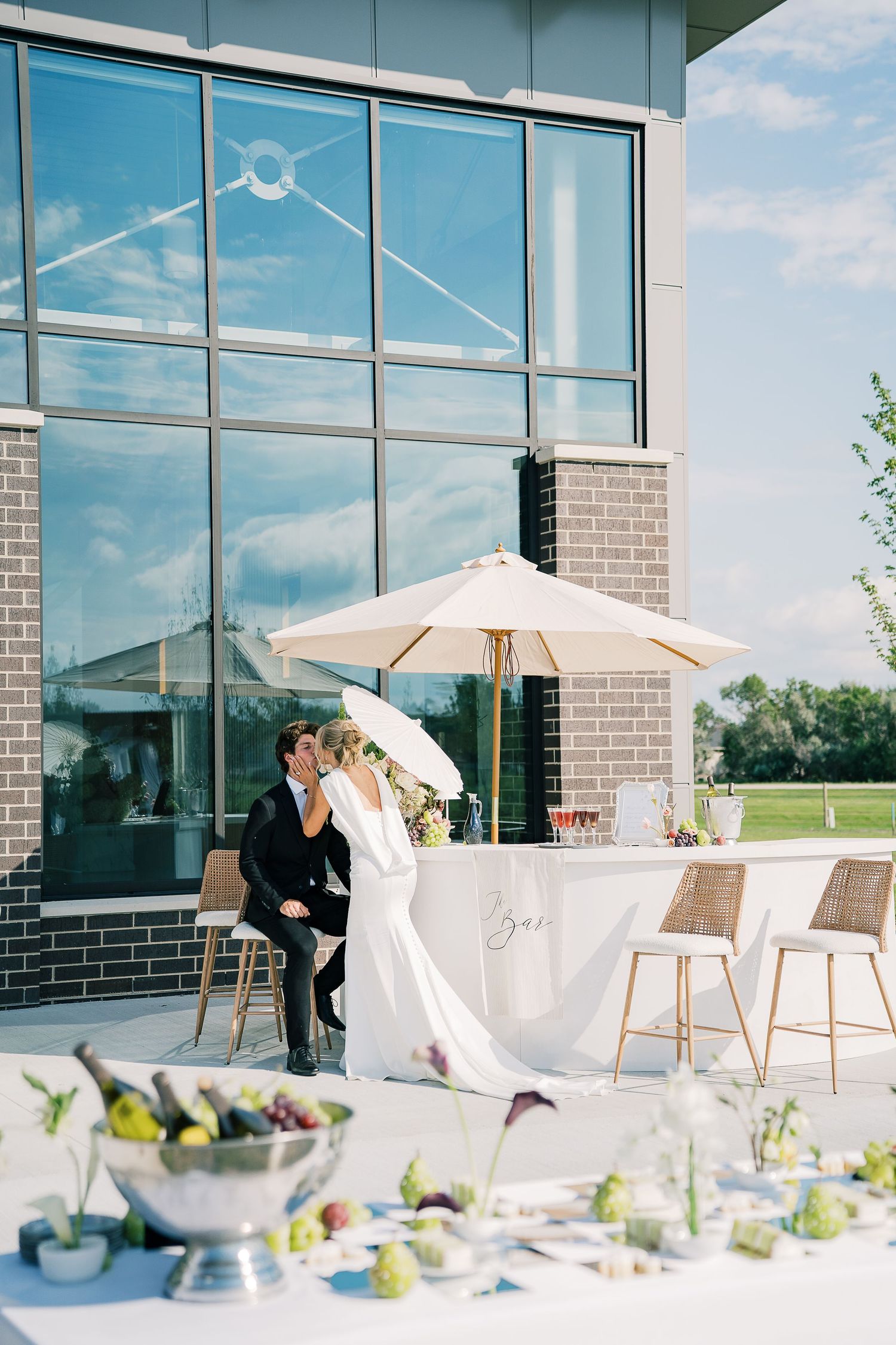 Elegant outdoor wedding reception setup with white tables, umbrellas and modern glass building backdrop on sunny day.