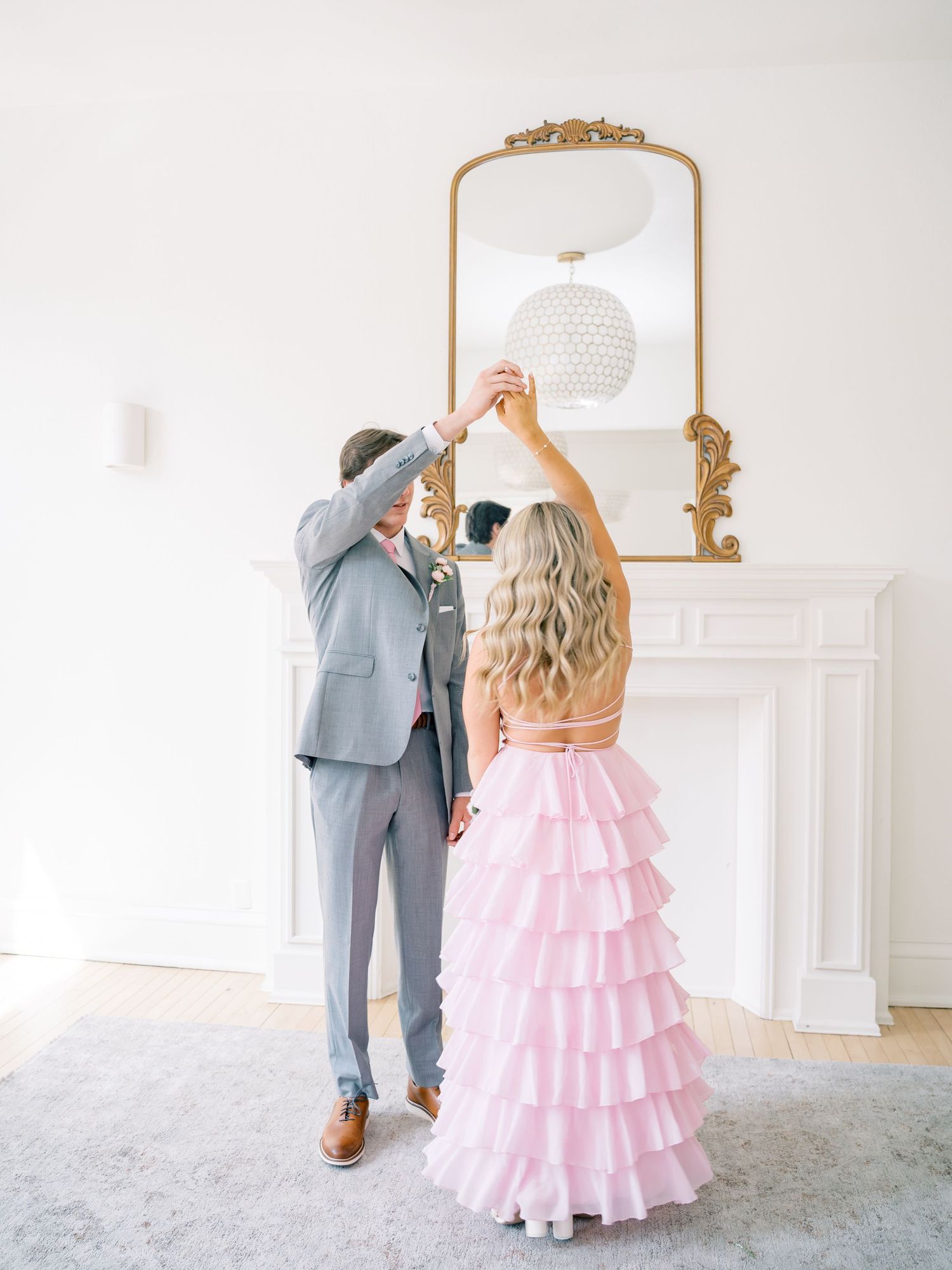 Couple dancing in elegant white room, woman in pink ruffled gown, man in gray suit, gold mirror above fireplace.