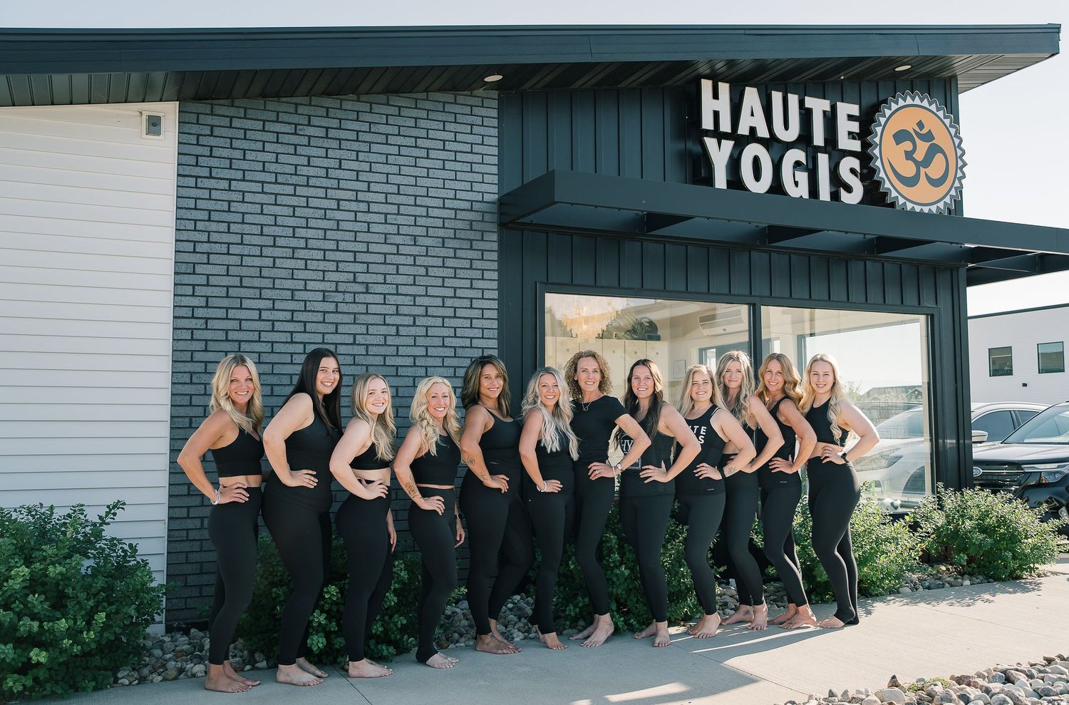 A group of yoga instructors wearing matching black outfits pose in front of Haute Yogis studio entrance.