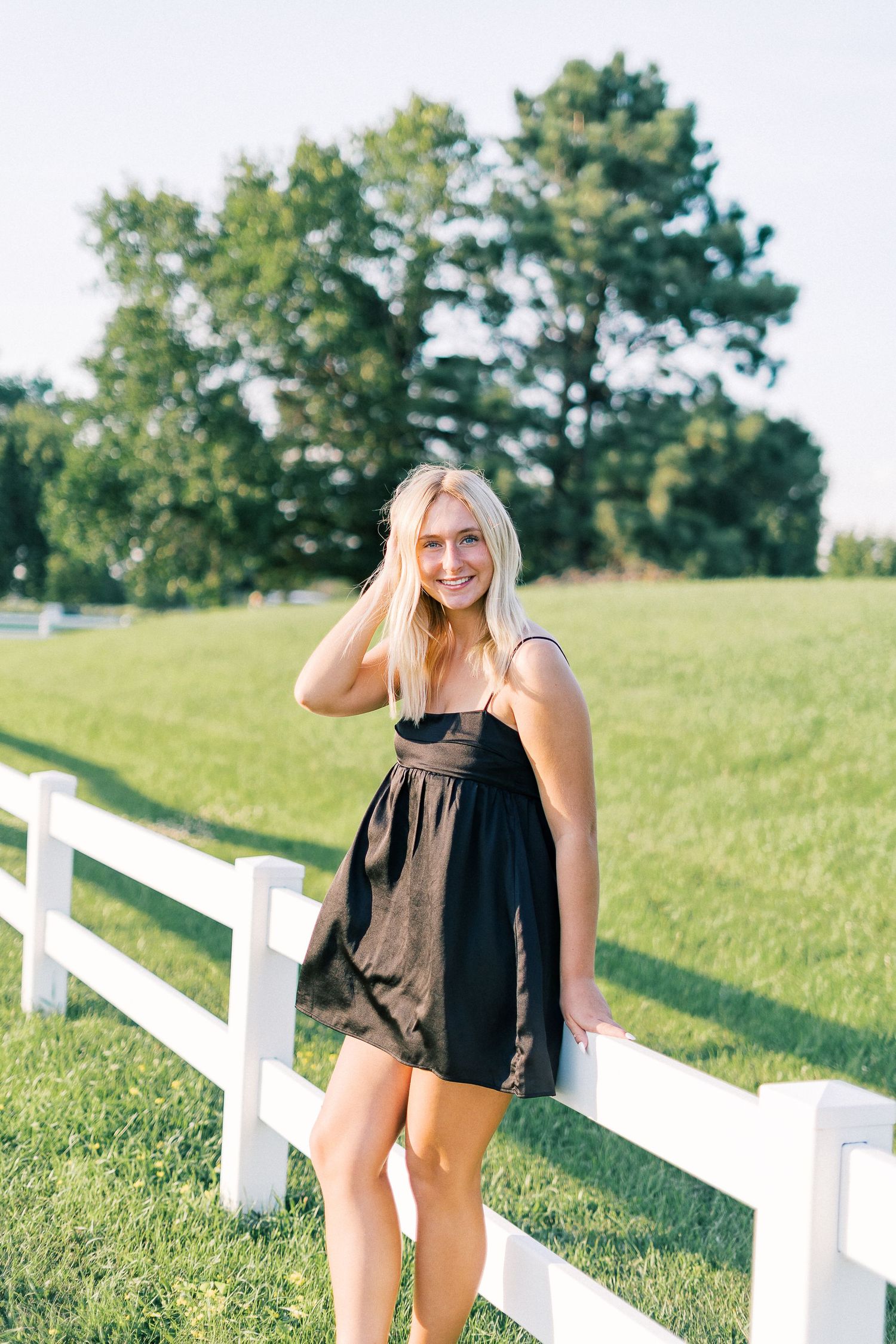 A young woman in a black sundress poses beside a white fence on a sunny summer day in a grassy field.