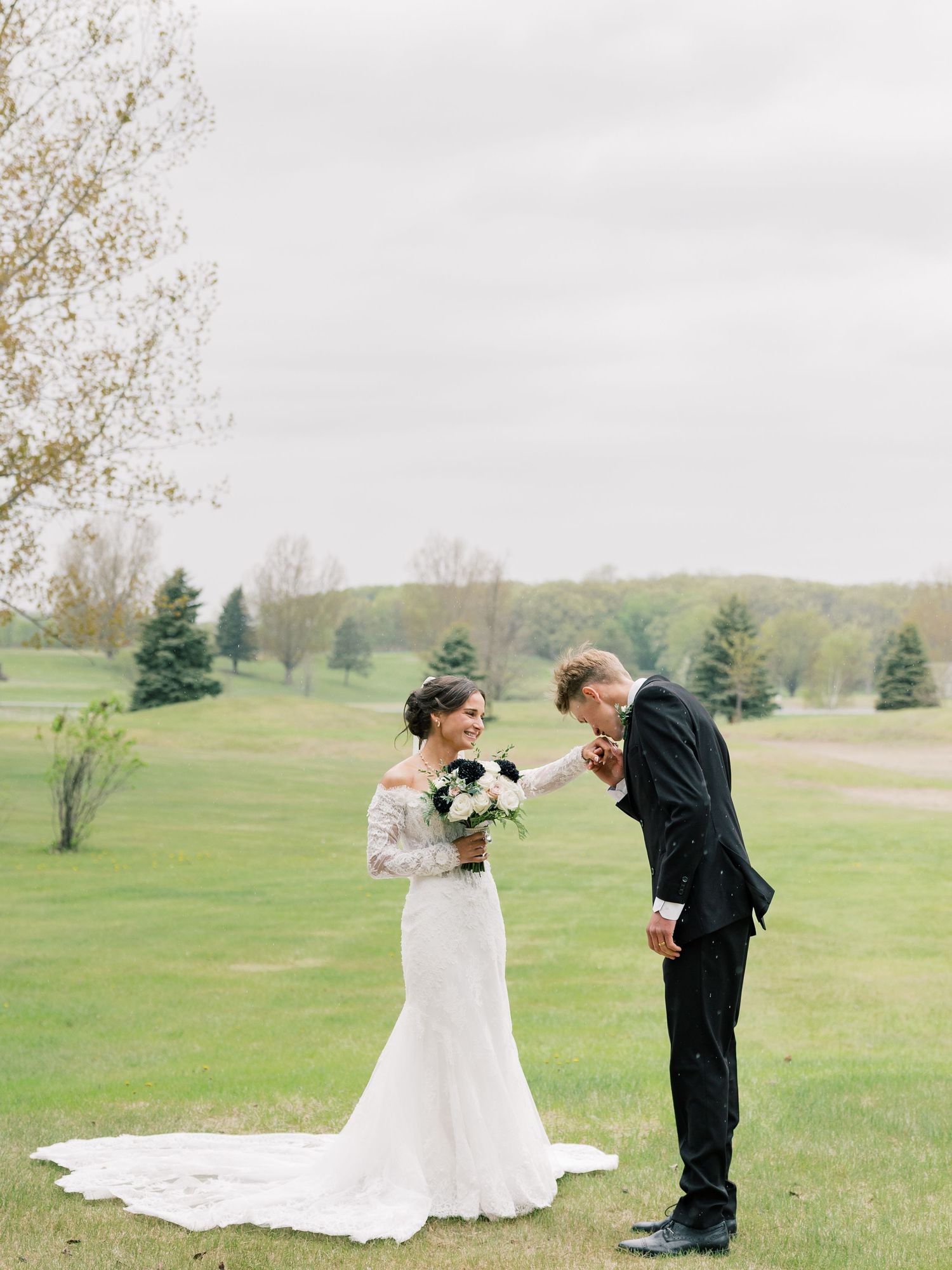 Groom kissing bride's hand in a lush green outdoor setting, bride wearing lace wedding gown holding black and white bouquet.