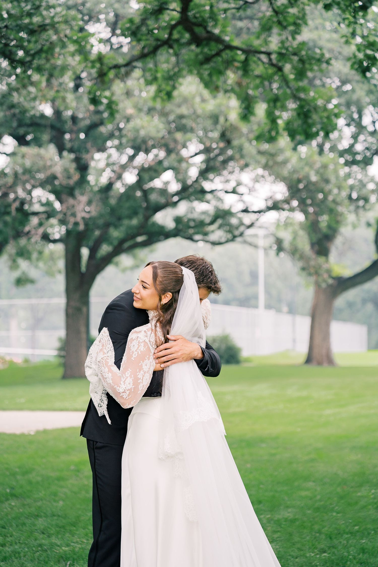 Bride and groom share a tender embrace outdoors, surrounded by lush green trees on their wedding day.